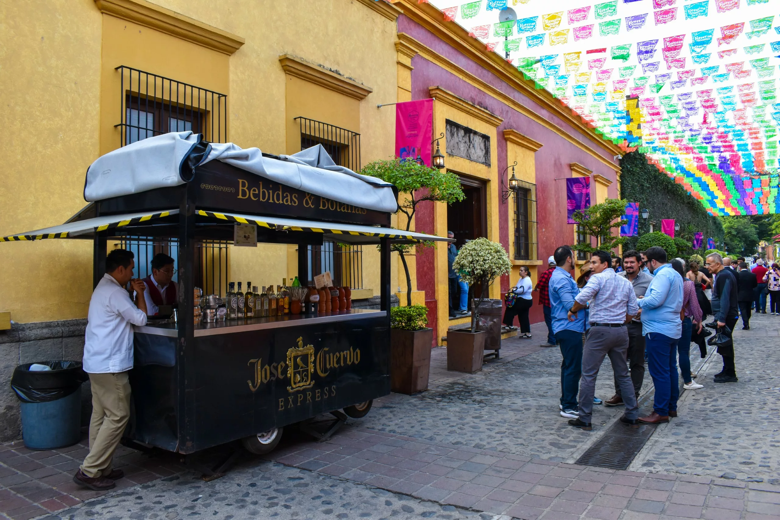 Une scène de rue animée dans la ville de Tequila, au Mexique, où les gens se pressent près d'un chariot noir de tequila « Jose Cuervo Express », sous des décorations festives en papel picado et devant des bâtiments aux couleurs vives.
