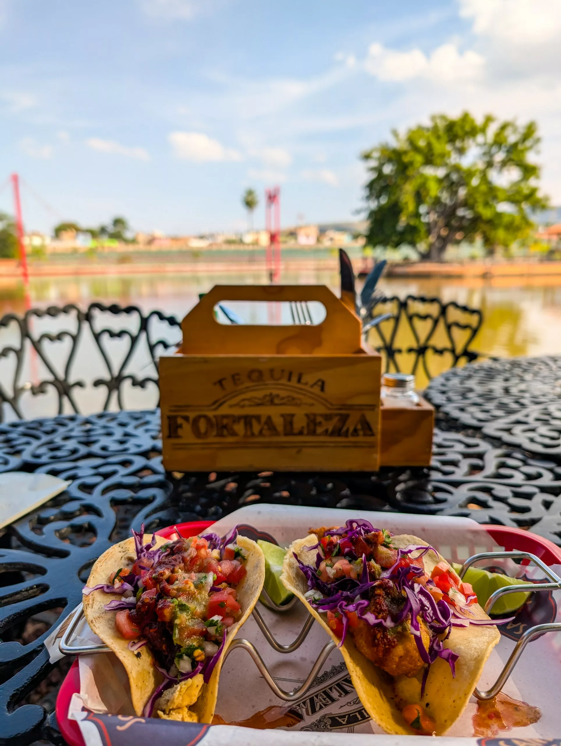 Deux tacos garnis de couleurs vives trônent sur une table en métal au bord d’une rivière, à côté d’une caisse en bois portant l’inscription « Tequila Fortaleza » .