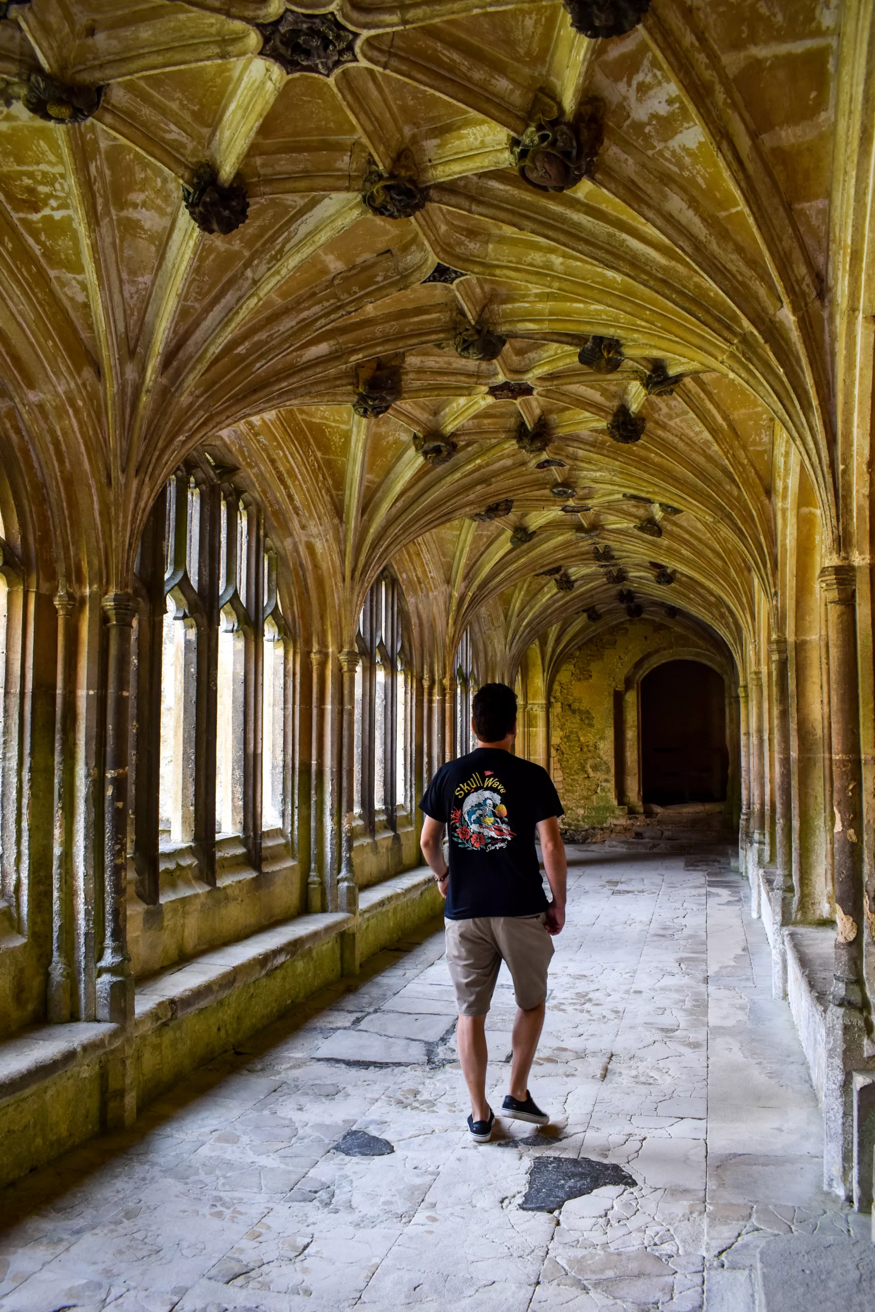 Une personne vêtue d'un t-shirt à motif et d'un short traverse un cloître en pierre au plafond voûté visible dans les films Harry Potter