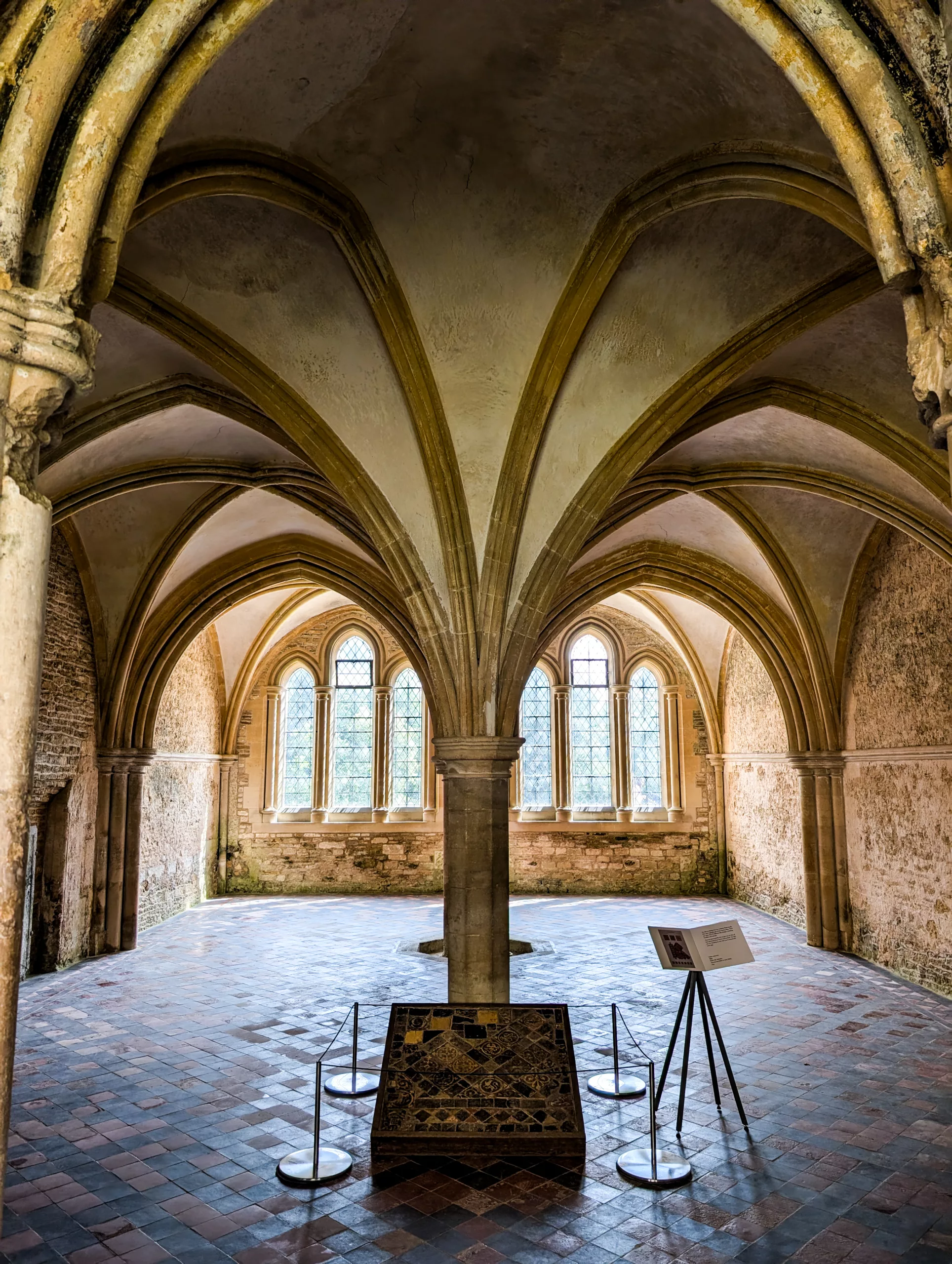 Intérieur d'une pièce en pierre avec des voûtes à nervures, une colonne centrale et un sol carrelé, rappelant les décors de Harry Potter, avec un petit présentoir au premier plan.