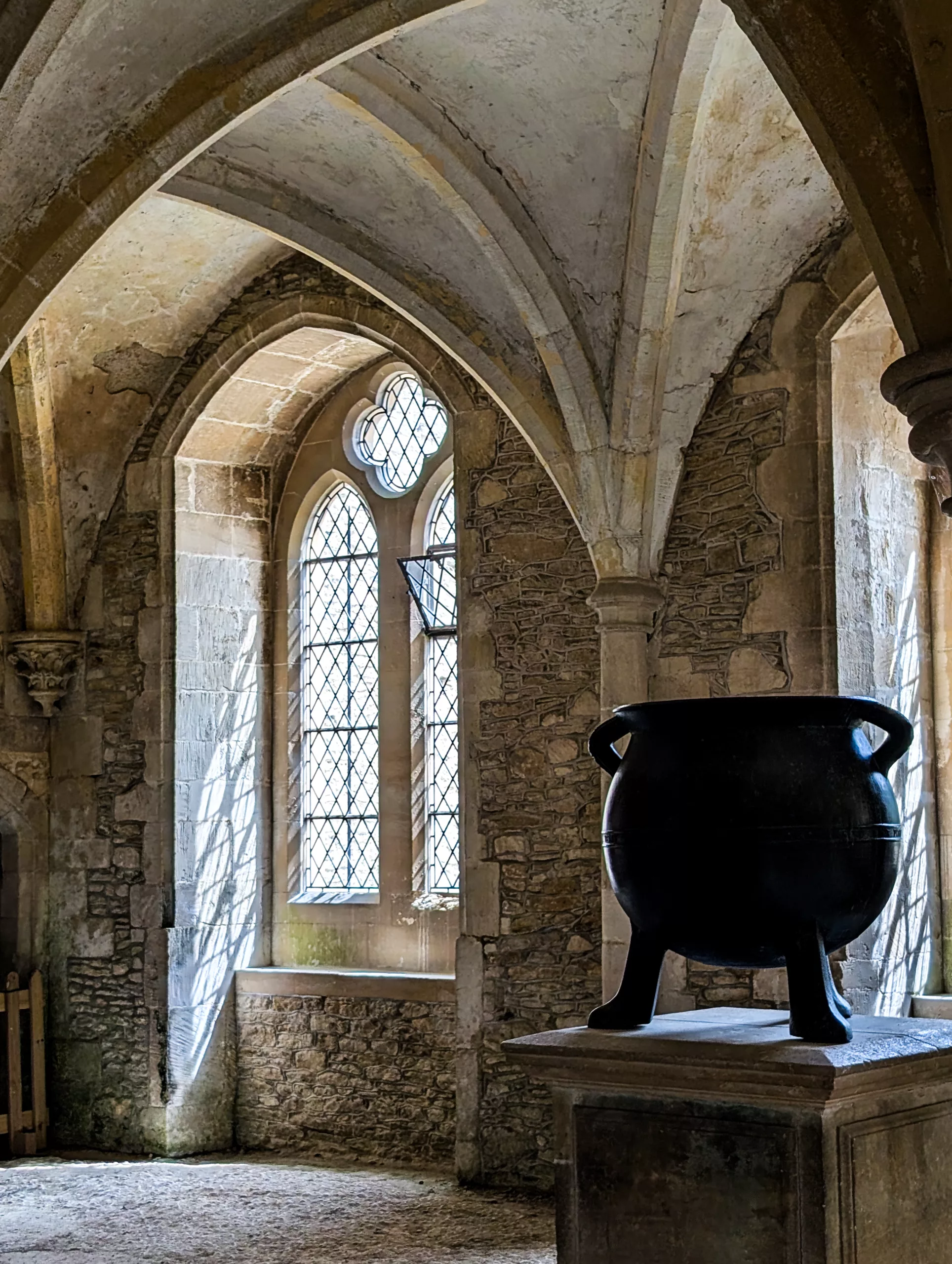 Un grand chaudron noir trône sur un socle en pierre au cœur d'une pièce aux murs de pierre, dotée d'un plafond voûté et d'une haute fenêtre cintrée qui rappelle l'univers d'Harry Potter.