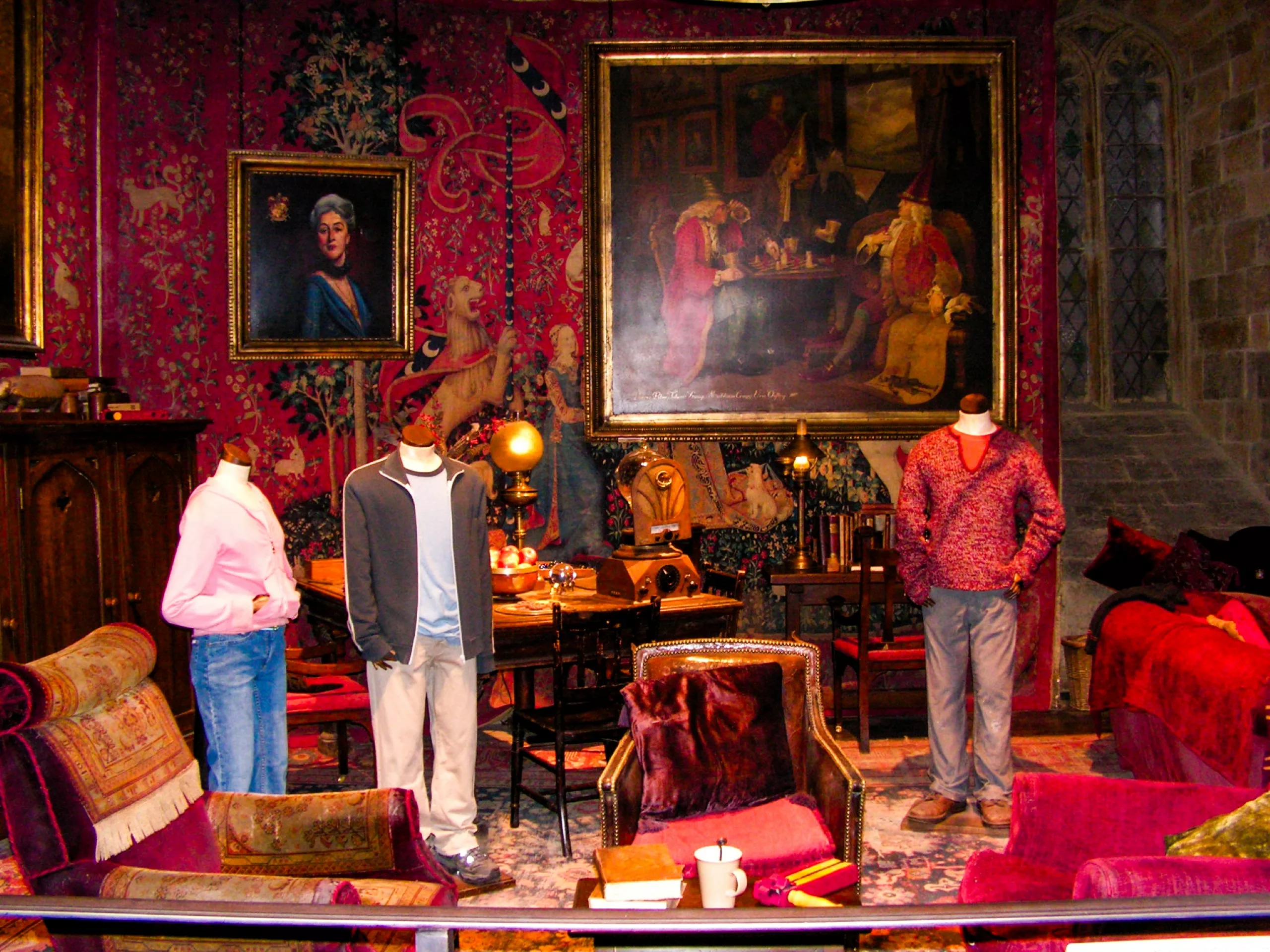 Trois mannequins représentant Harry, Ron et Hermione, vêtus de tenues décontractées se tiennent dans une pièce décorée d'une tapisserie florale rouge, de tableaux encadrés et de meubles anciens, représentant la salle commune de Gryffondor