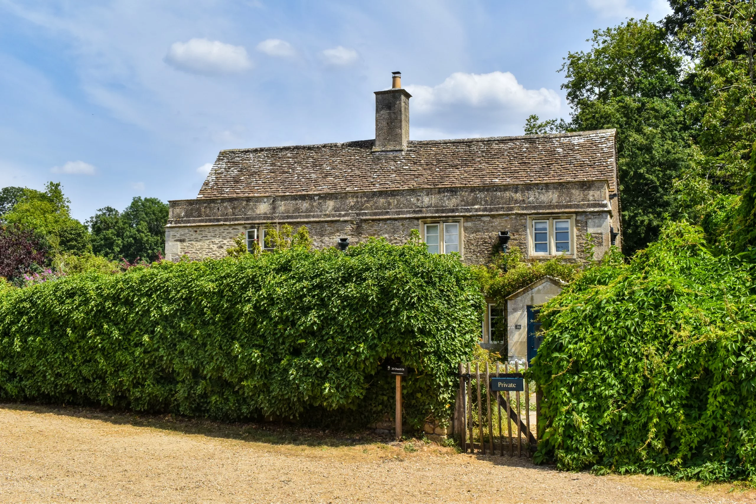 Une maison en pierre à deux étages, avec un toit en ardoise, nichée derrière une haute haie et un portail en bois portant l'inscription « Privé ».