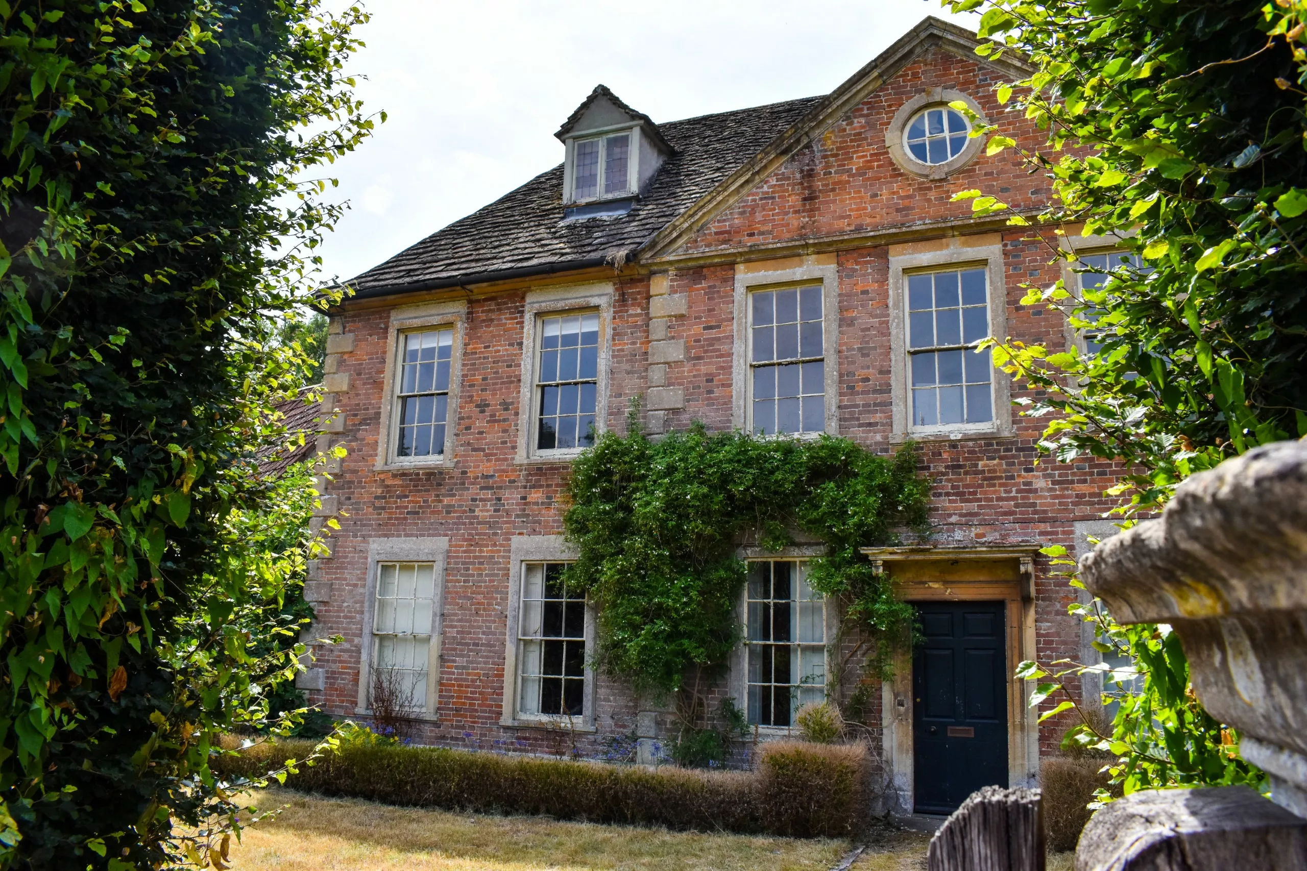Une maison en briques de deux étages, avec un toit à pignon, des fenêtres à guillotine et une porte d'entrée sombre, partiellement encadrée par de la verdure