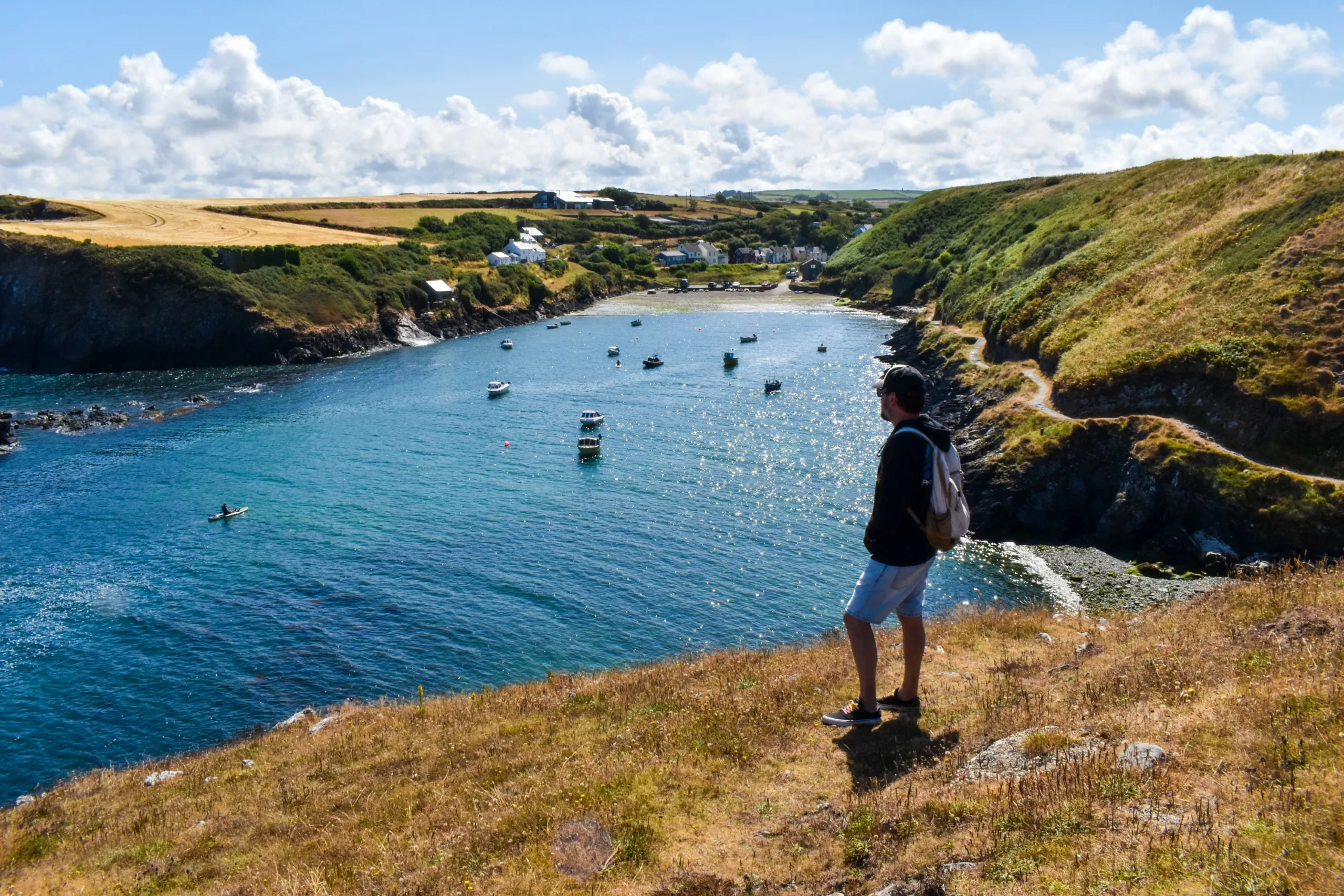 Une personne avec un sac à dos se tient sur une falaise, surplombant une baie côtière au Pays de Galles, où de petits bateaux et un kayakiste dérivent en contrebas.