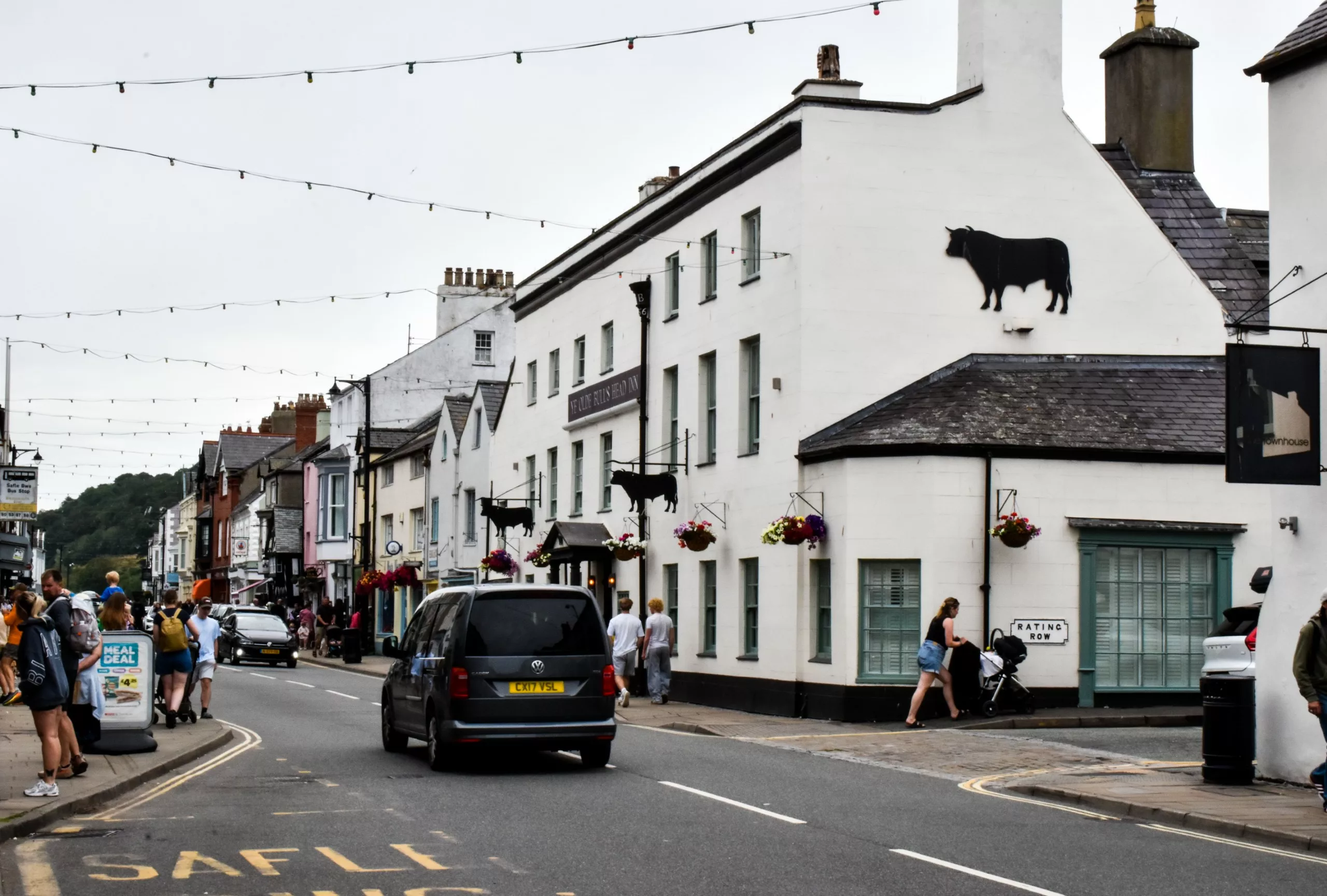 Des passants passent devant l'auberge « Ye Olde Bulls Head Inn » dans une ville galloise tandis qu'un monospace sombre passe devant eux ; le bâtiment blanc est orné d'une grande silhouette noire représentant un taureau