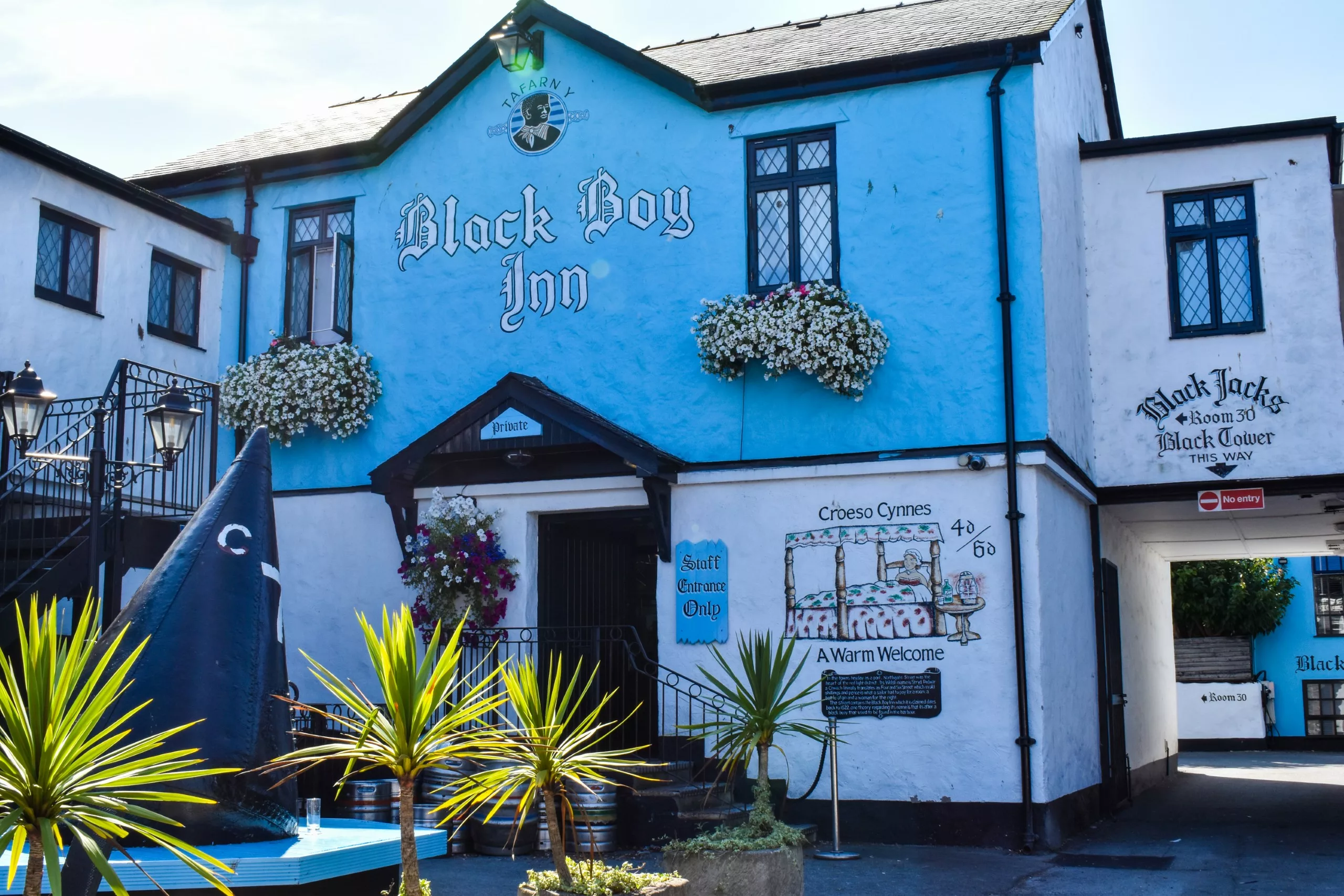 La façade bleue du Black Boy Inn. Il y a une fresque représentant un lit à baldaquin, des jardinières fleuries et des panneaux indiquant aux clients des directions.