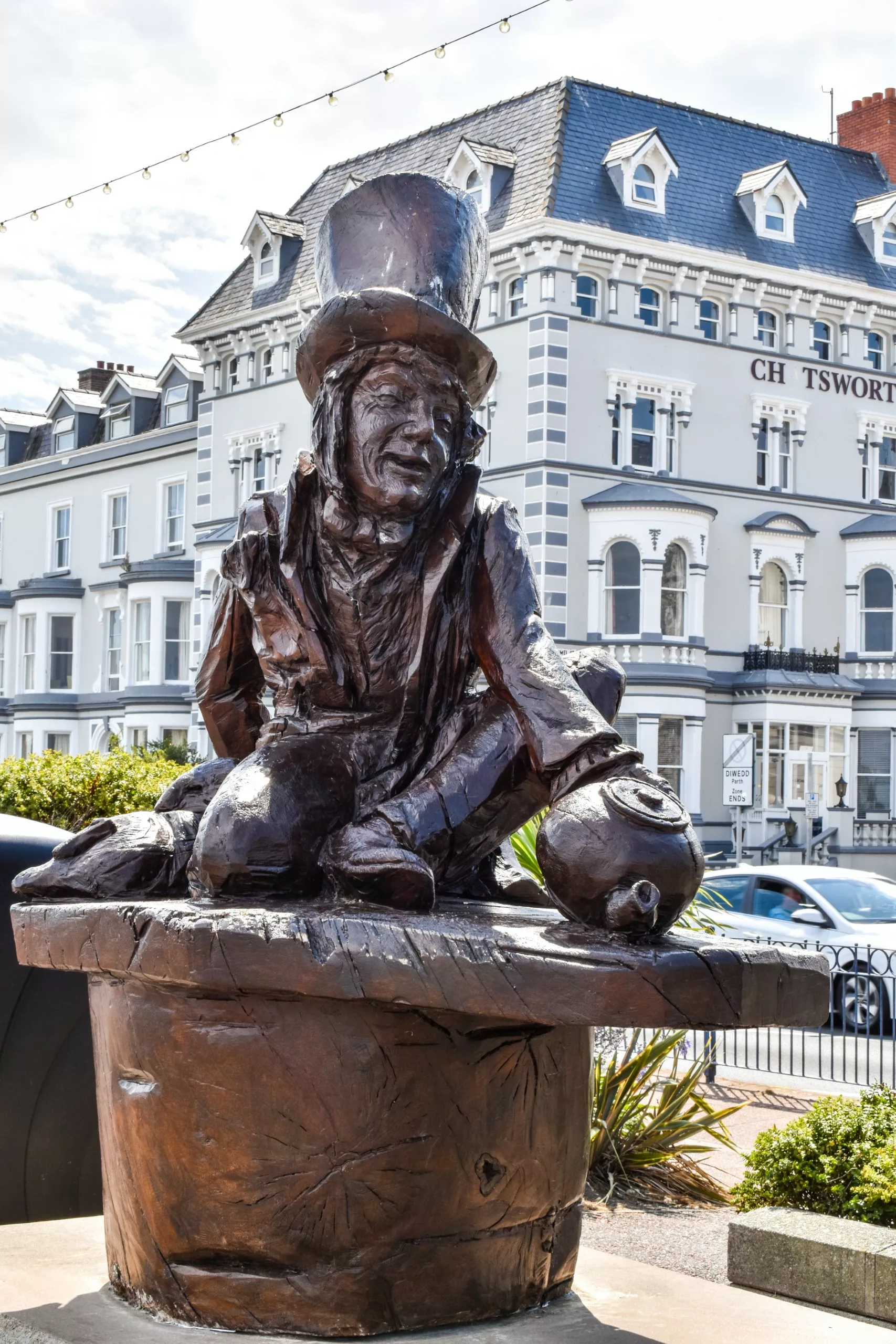 Une sculpture en bronze représentant le Chapelier fou d'Alice au Pays des Merveilles agenouillé sur une souche d'arbre, avec en arrière-plan un grand bâtiment de style victorien