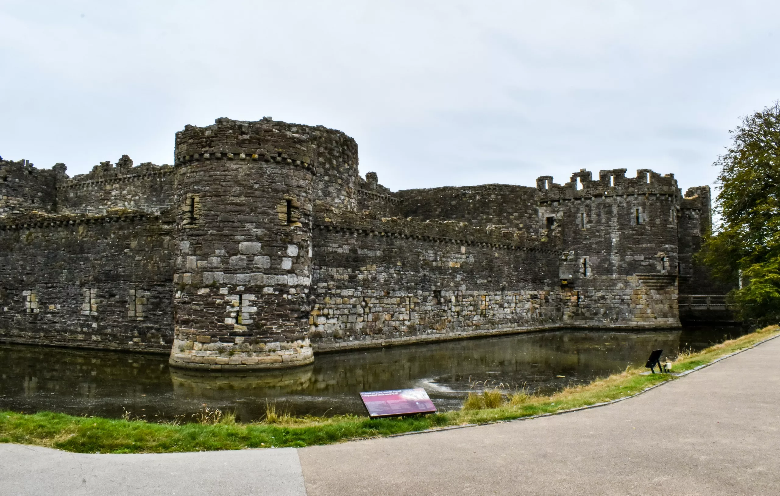 Les murs de pierre et la tour circulaire d'un château médiéval entouré d'un fossé rempli d'eau, avec au premier plan un chemin pavé