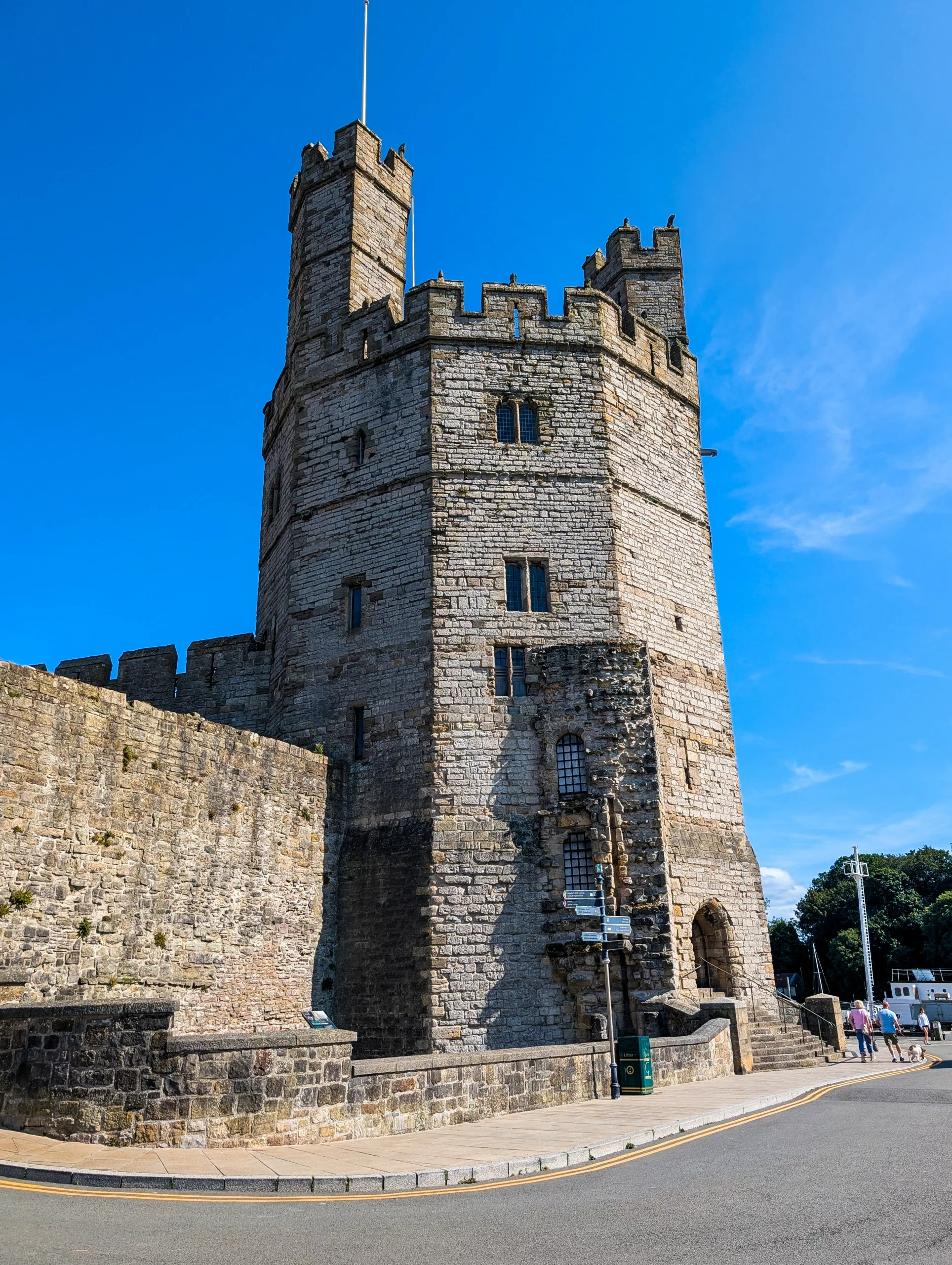 Une haute tour de château en pierre à plusieurs étages, dotée de remparts crénelés et d'un mur défensif, vue depuis un coin de rue sous un ciel bleu limpide