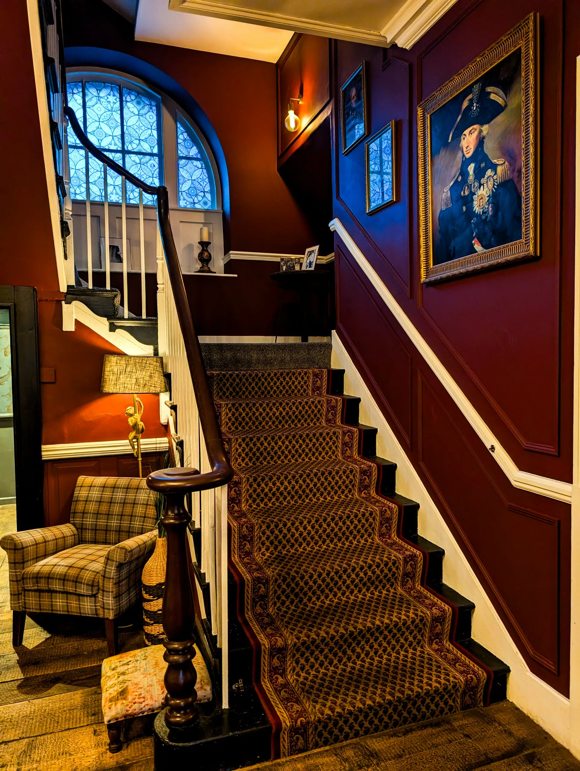 Un escalier orné d'un tapis à motifs, adossé à des murs lambrissés d'un rouge foncé décorés de portraits encadrés, à côté d'un fauteuil à carreaux et d'une grande fenêtre cintrée.