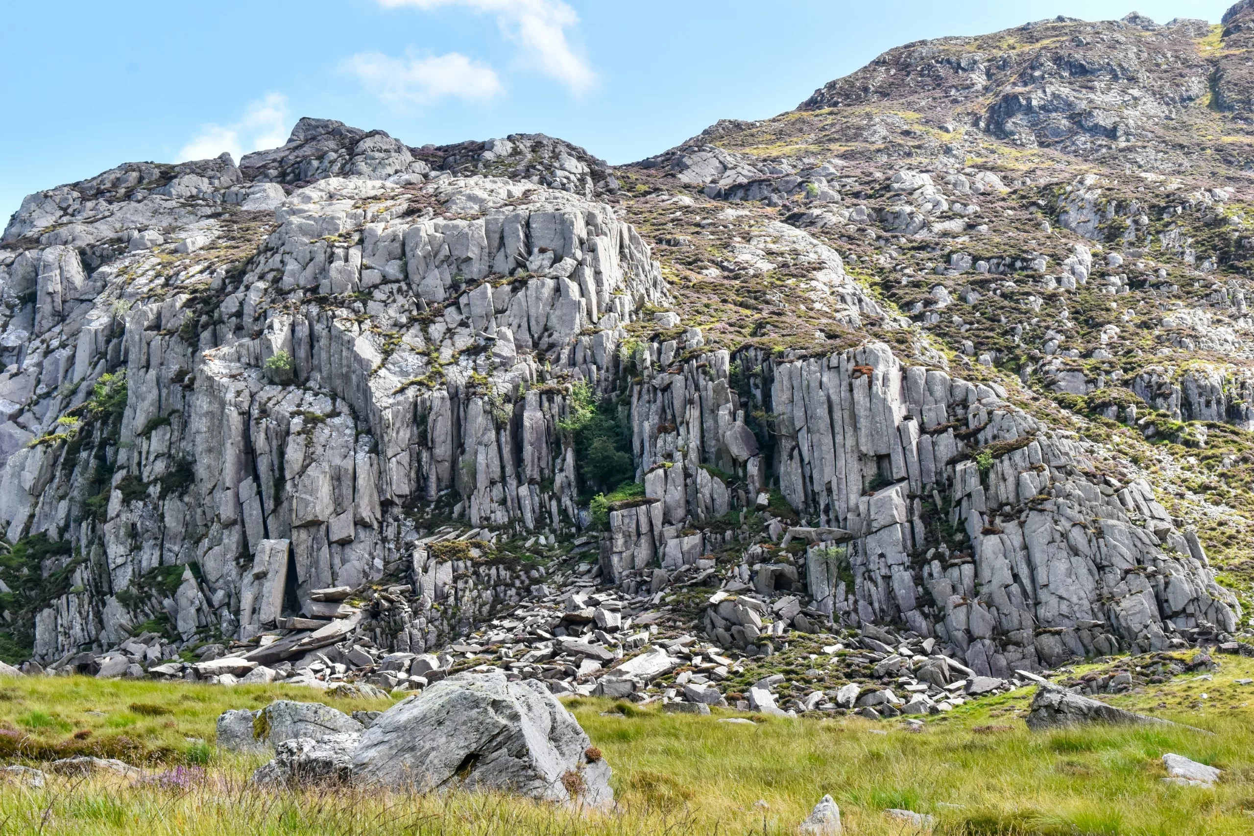 Une pente montagneuse escarpée et accidentée, avec des parois rocheuses verticales et un champ de rochers instables à sa base, qui s'élève au-dessus d'une prairie verdoyante, ce qui en fait une destination très prisée des visiteurs du Pays de Galles.