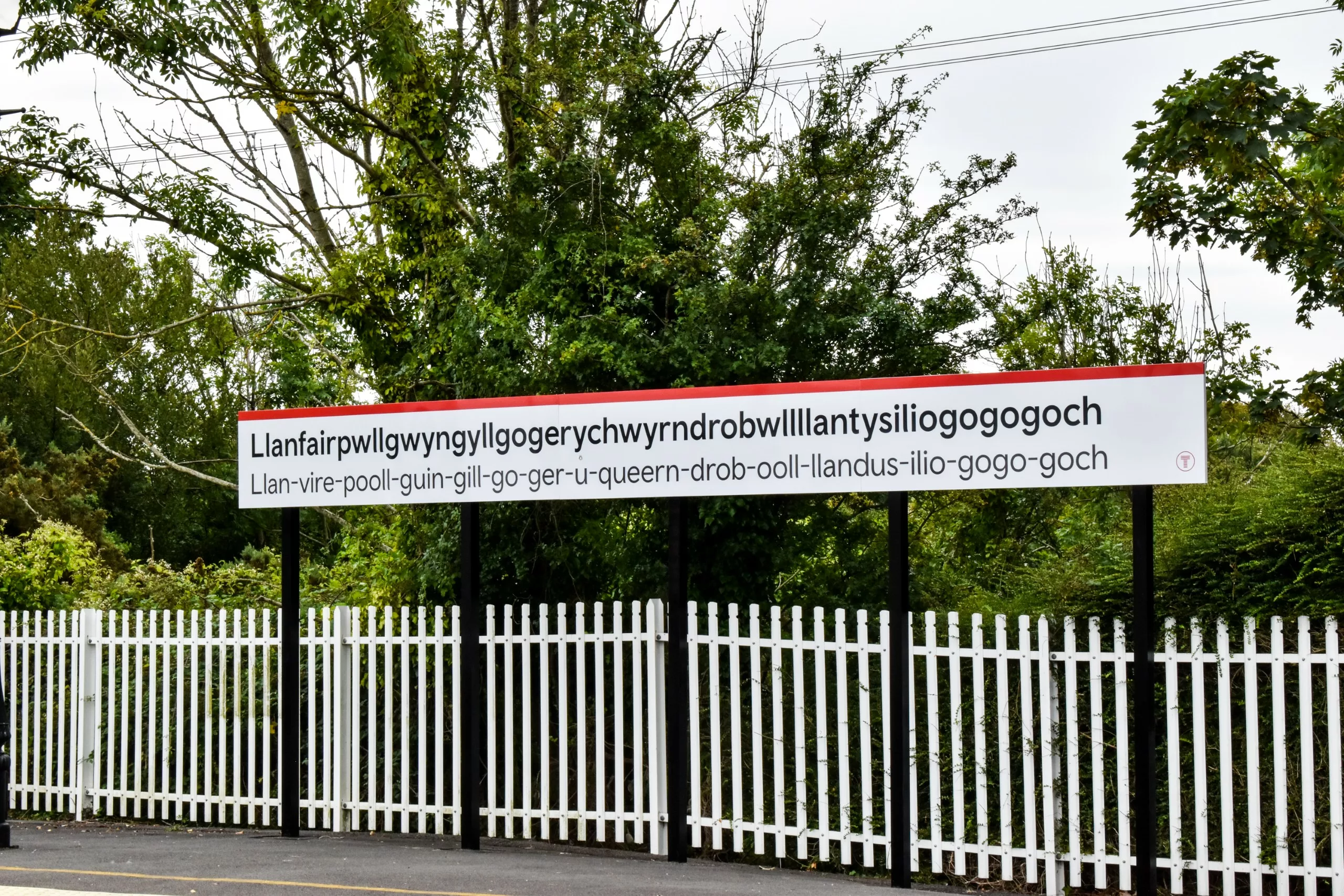 Un panneau blanc de gare, lieu très visité au Pays de Galles, sur lequel figure le nom « Llanfairpwllgwyngyllgogerychwyrndrobwllllantysiliogogogoch » en gallois, accompagné de sa transcription phonétique.