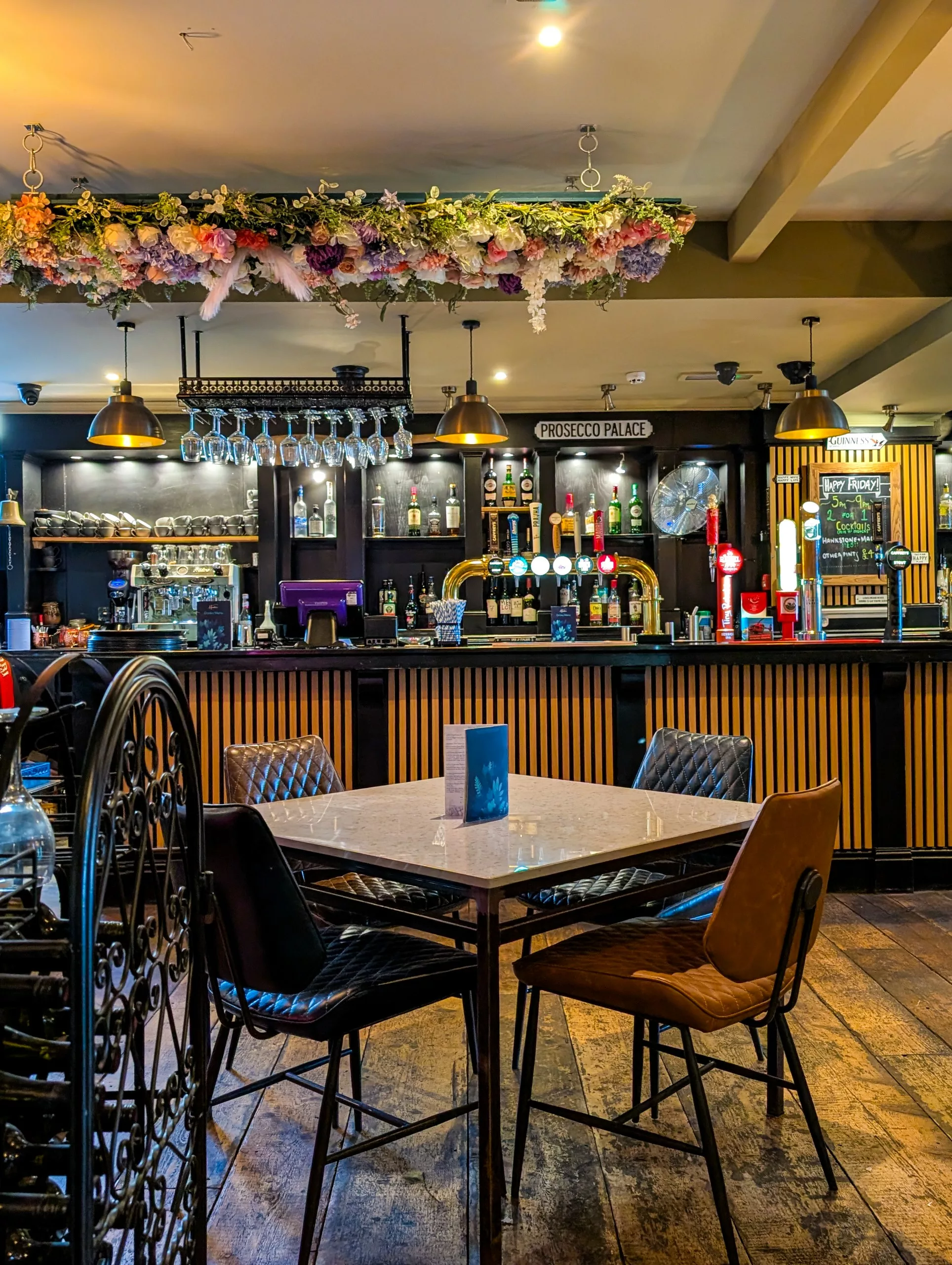 Une table carrée entourée de quatre chaises est disposée devant un long comptoir orné d'une composition florale suspendue, de robinets à bière et d'étagères remplies de bouteilles d'alcool.