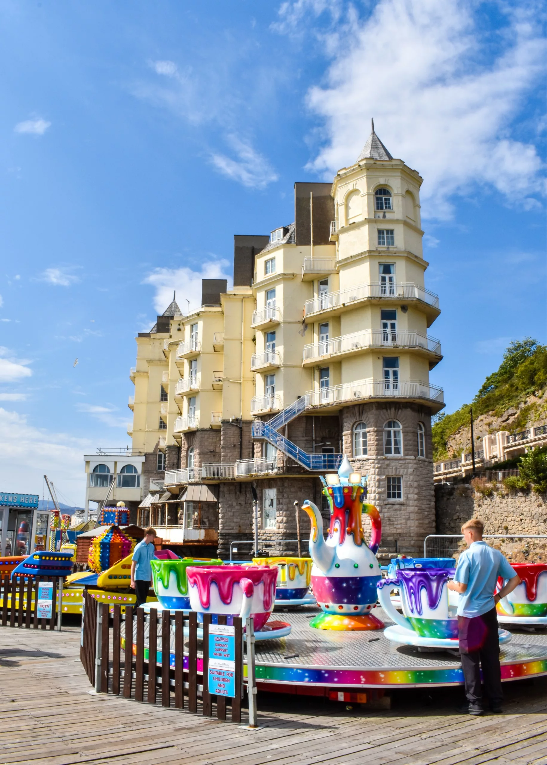 Un manège de tasses tournantes sur une jetée en bois, devant un grand immeuble jaune de plusieurs étages à Llandudno