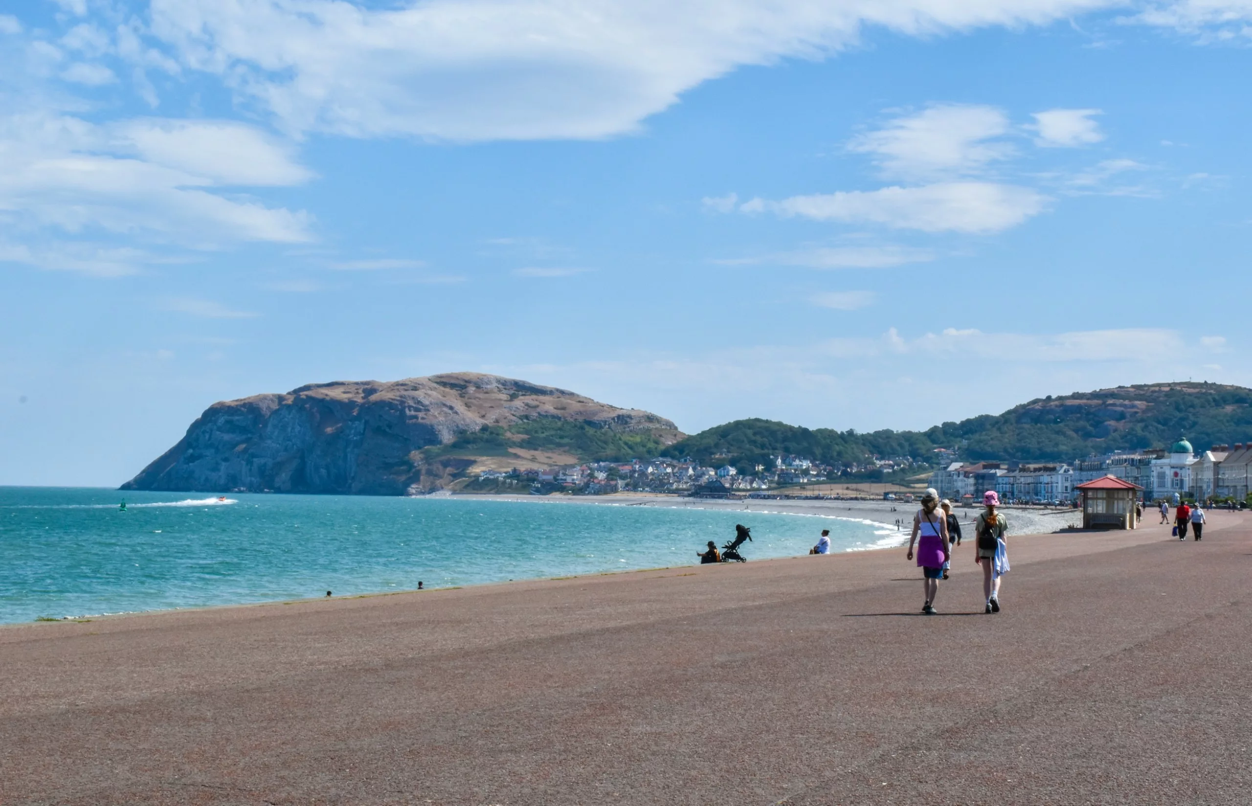 Des promeneurs déambulent le long d'une promenade pavée au bord de l'océan, avec un grand promontoire en arrière-plan