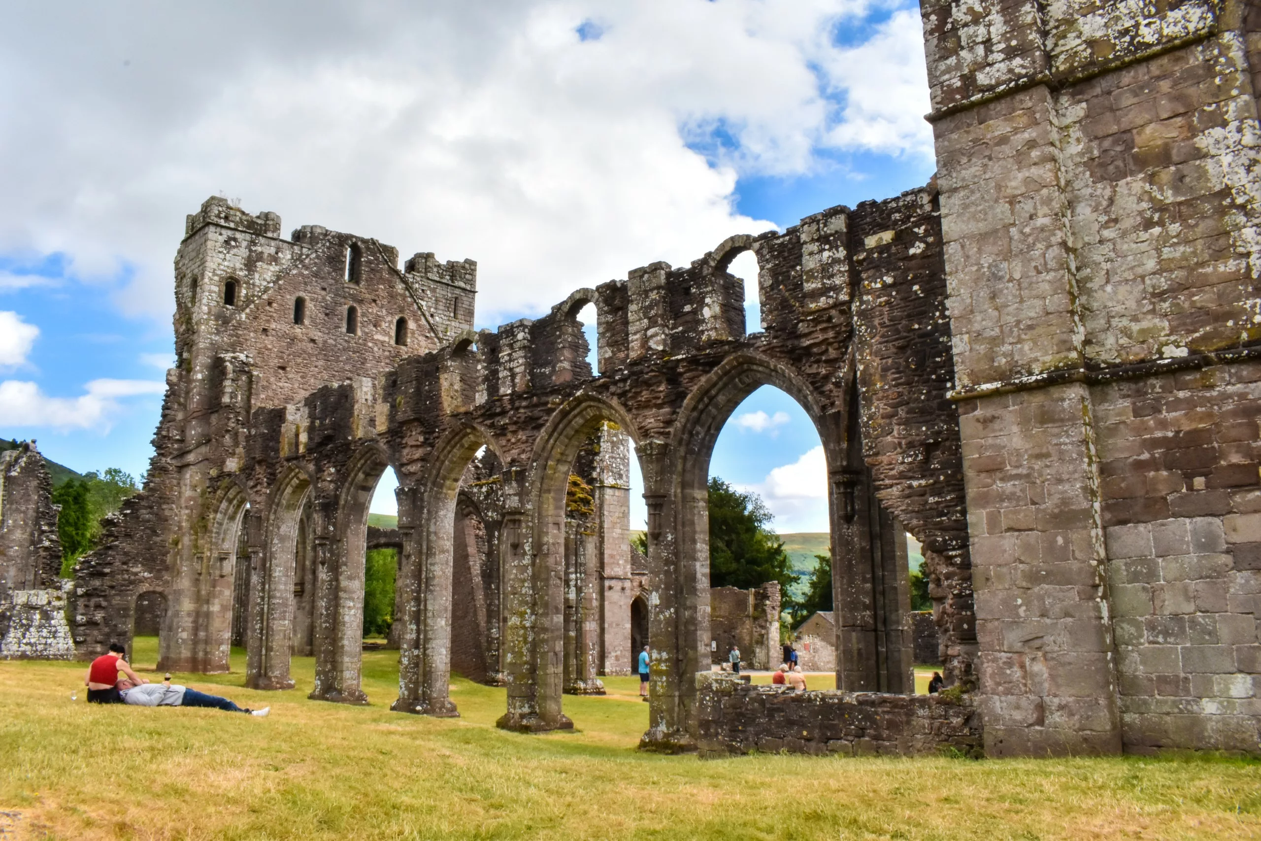 Ruines en pierre d'une abbaye médiévale au Pays de Galles, avec de hautes colonnes cintrées, situées dans un champ d'herbe où se trouvent plusieurs visiteurs assis.