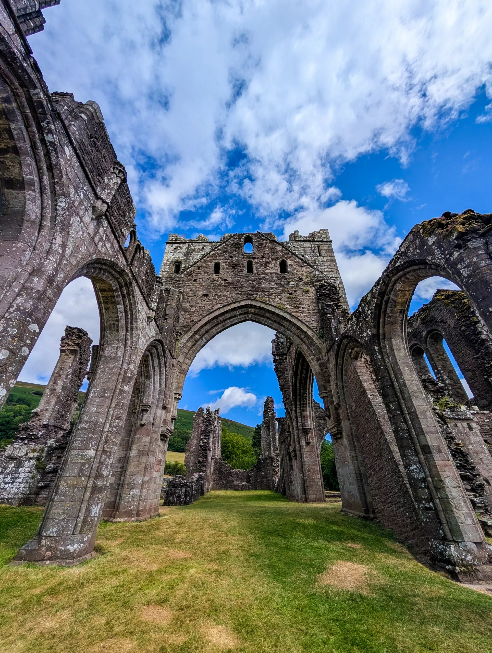 Intérieur de ruines en pierre d'une abbaye gothique au Pays de Galles, avec de grandes portes voûtées sous un ciel bleu parsemé de nuages.