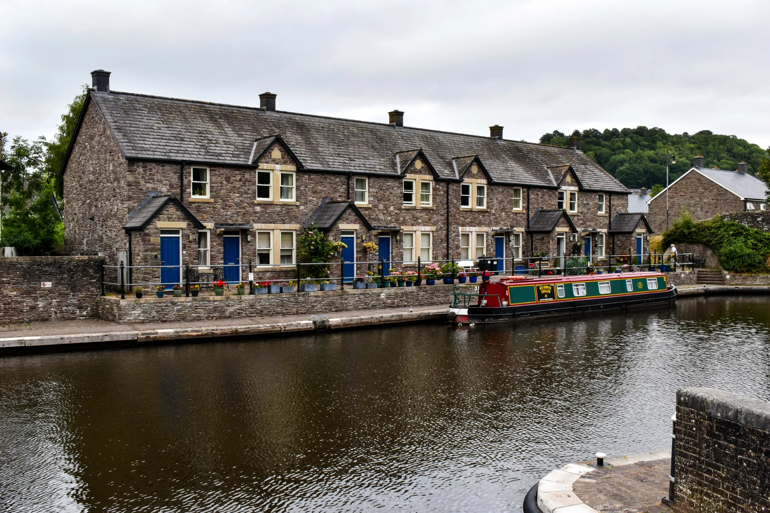 Une rangée de petites maisons en pierre aux portes bleues longe un canal, devant lequel est amarré un long bateau étroit dans la ville de Brecon.