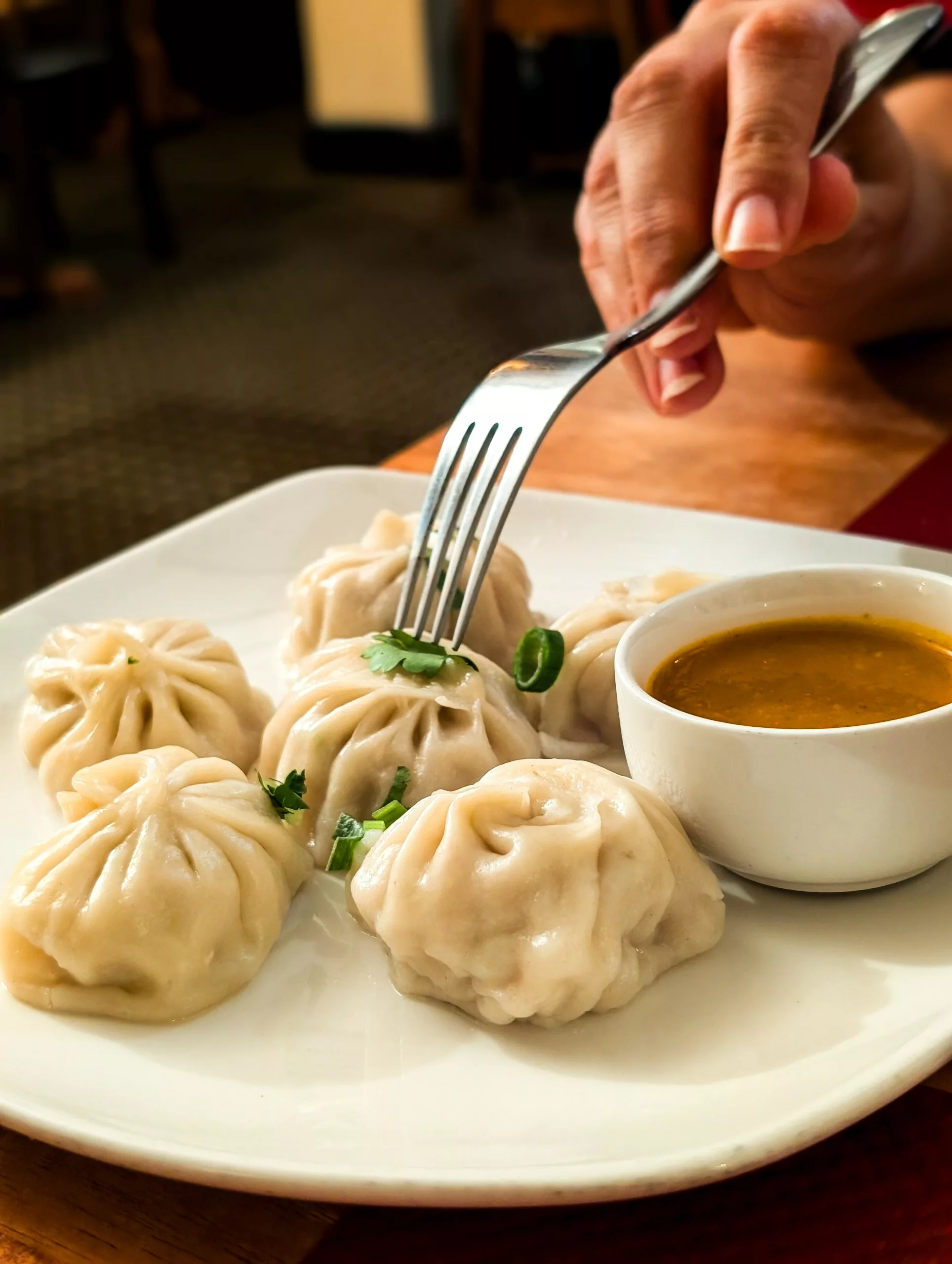 Une main utilise une fourchette pour prendre un raviolis en boule cuit à la vapeur dans une assiette qui en contient cinq. A côté, se trouve une sauce dans laquelle on trempe les boulettes.