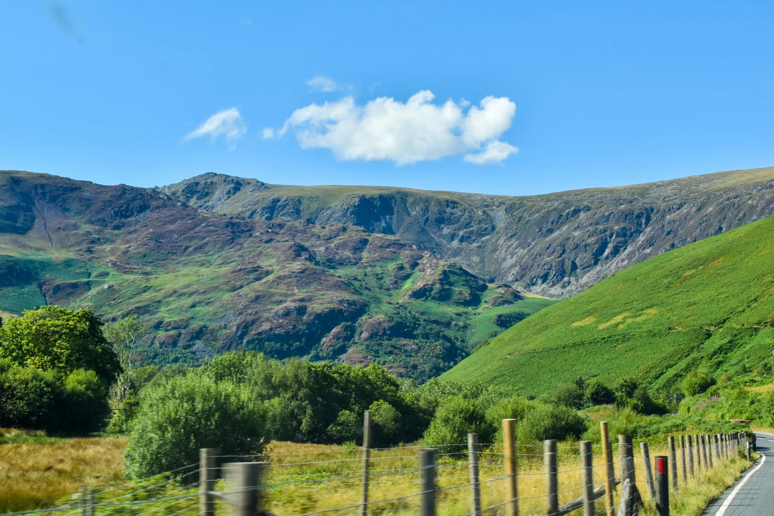 Une route longe une clôture métallique, avec en toile de fond des collines verdoyantes et des montagnes escarpées et rocheuses, sous un ciel bleu limpide.