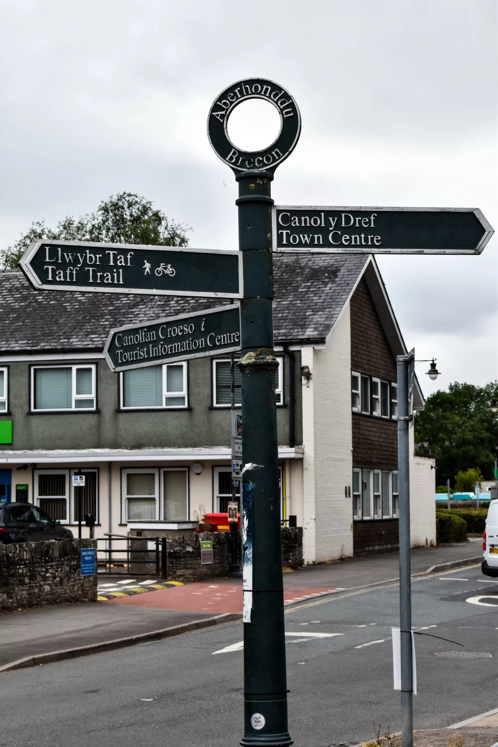 Un panneau indicateur métallique noir surmonté d'une inscription « Aberhonddu Brecon », indiquant la direction du centre-ville, du Taff Trail et de l'office de tourisme pour les visiteurs du Pays de Galles.