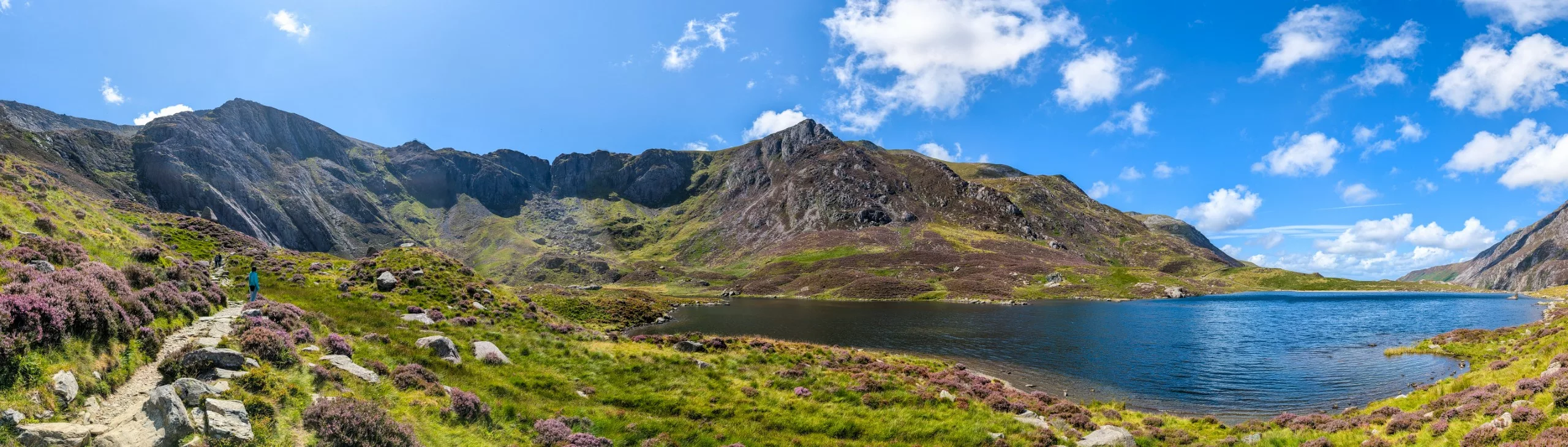 Une personne marche sur un sentier de pierre au bord d'un lac de montagne aux eaux sombres, au Pays de Galles, entourée de collines recouvertes de bruyère violette et de pics rocheux, sous un ciel bleu.