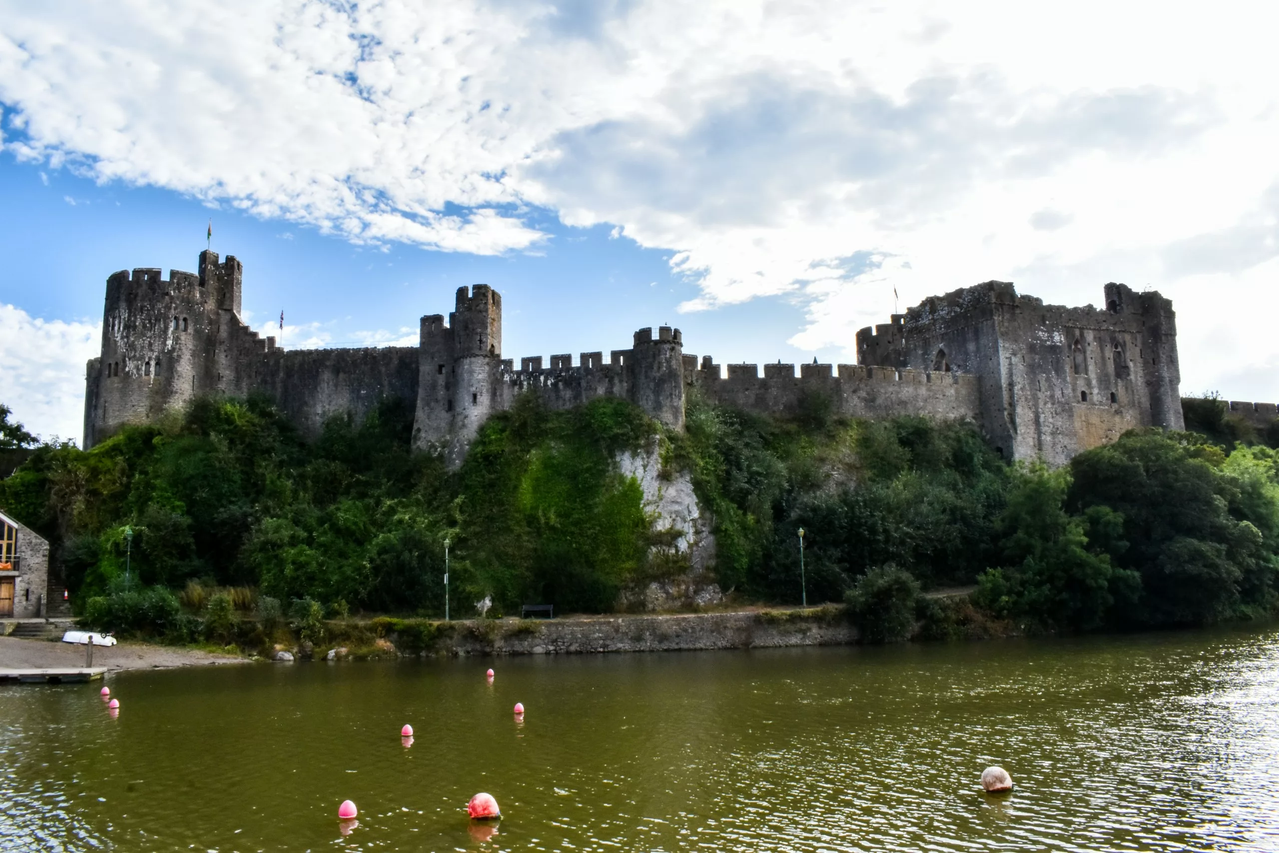 Un château en pierre doté de plusieurs tours se dresse au sommet d'une falaise recouverte d'herbe surplombant une rivière ; c'est le château de Pembroke, à visiter au Pays de Galles.