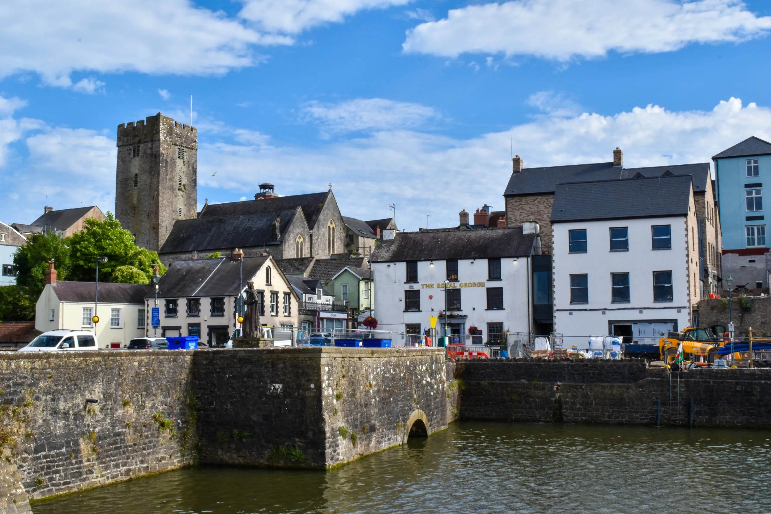 Un front de mer bordé de murs et de bâtiments en pierre, parmi lesquels le pub « The Royal George » et le haut clocher en pierre d'une église, au Pays de Galles.