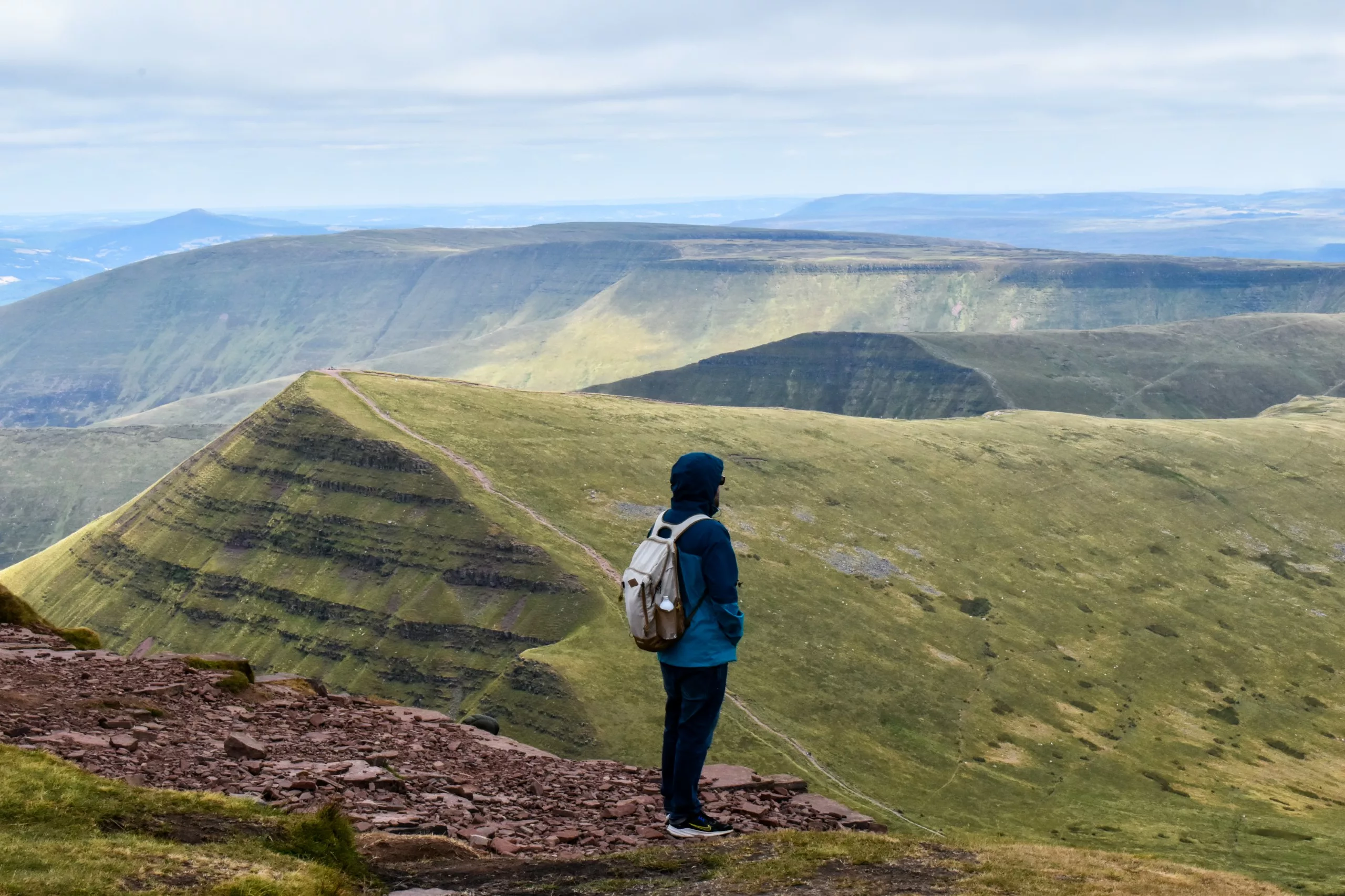 Un randonneur, sac à dos sur le dos, se tient sur une crête de la montagne Pen-y-Fan au Pays de Galles, d'où il contemple un vaste paysage vallonné et vert.
