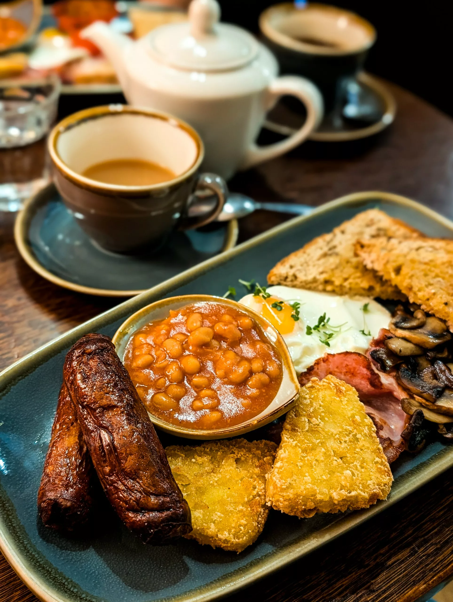 Petit déjeuner typique anglais/gallois avec une assiette de saucisses, œufs, bacon, champignons, haricots blancs à la tomate, toasts et rostis de patates. Une tasse de thé au lait est visible au second plan.