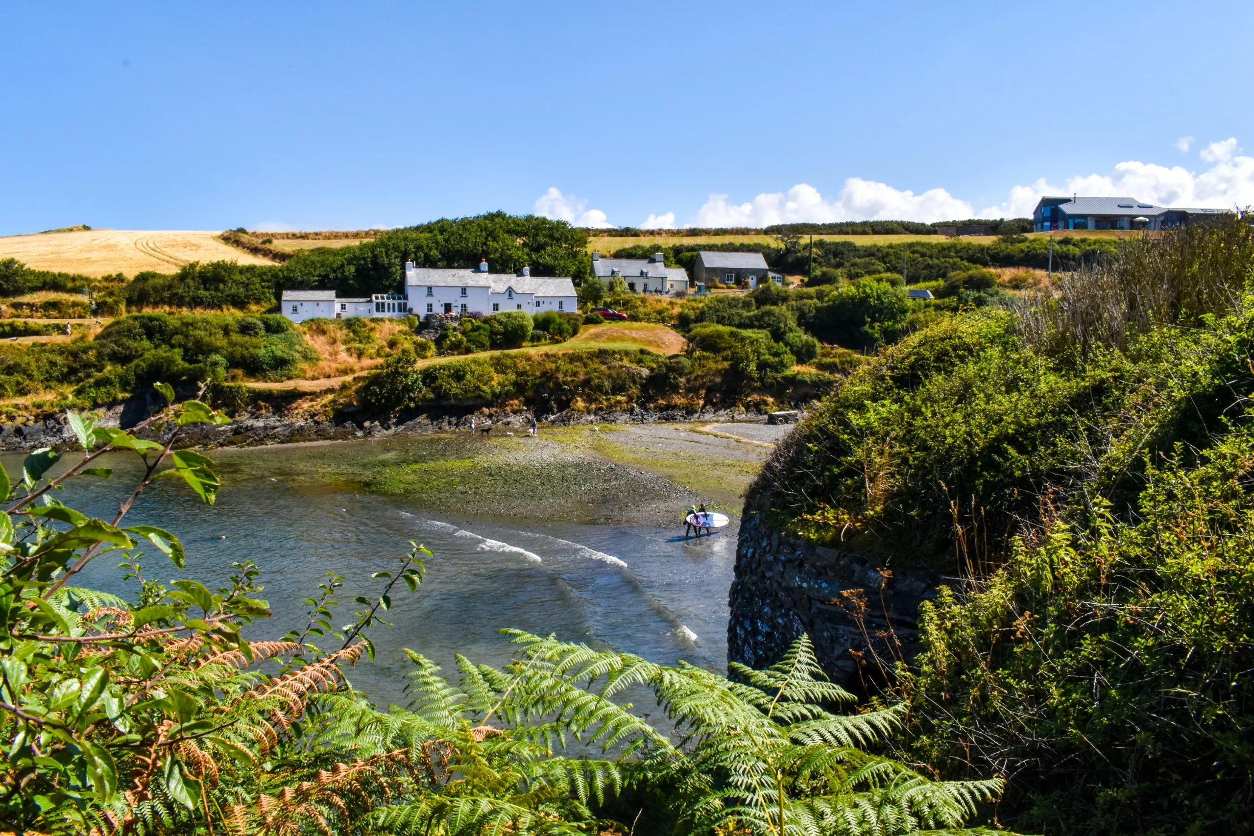 Deux personnes portent leurs planches de surf jusqu'à l'eau dans une crique, où une petite plage s'étend au pied de maisons blanches à flanc de colline.