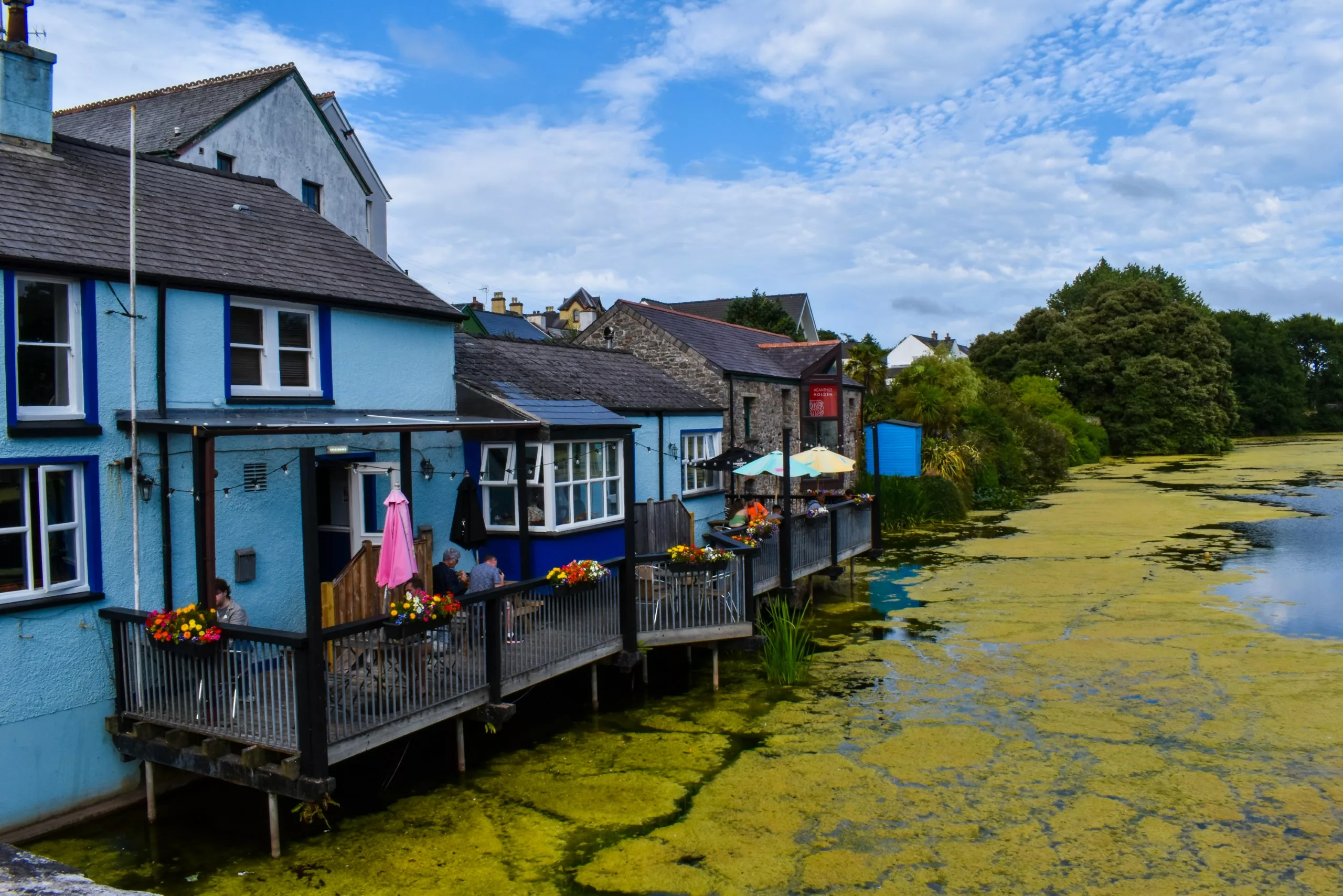 Des bâtiments bleus dotés de terrasses au bord de l'eau et de tables en plein air, le long d'un canal recouvert d'algues