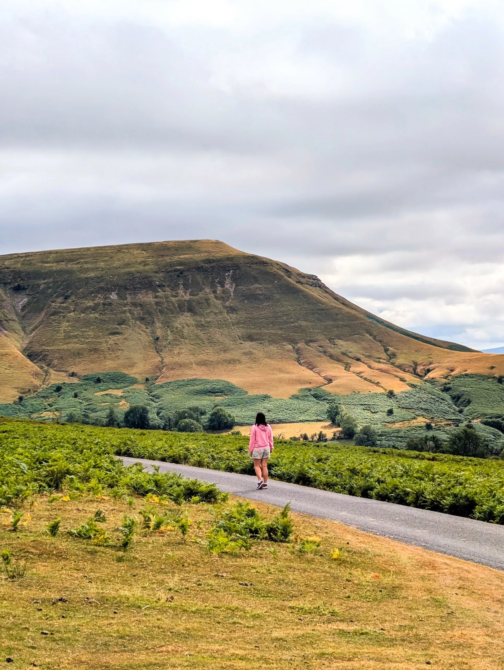 Une personne vêtue d'un sweat à capuche rose marche le long d'une route goudronnée menant à une grande montagne verdoyante au Pays de Galles, sous un ciel nuageux.
