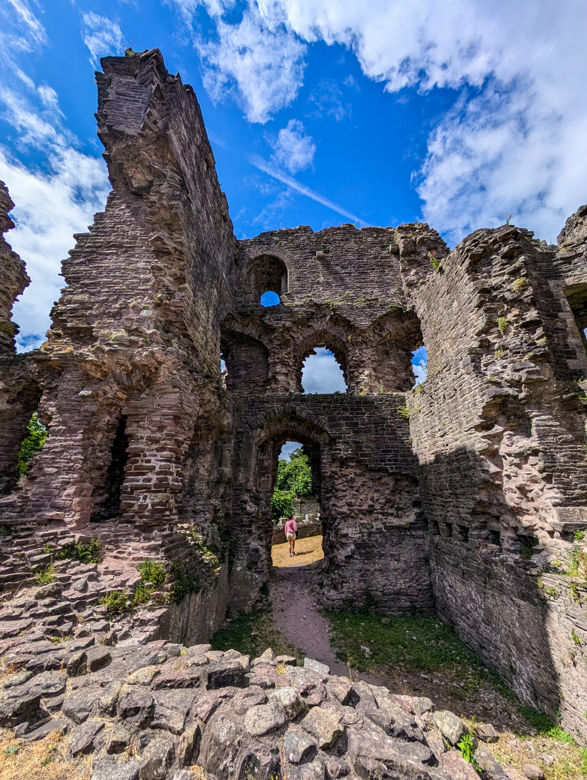 Un visiteur se tient à l'intérieur des ruines en pierre d'un château au Pays de Galles, devant des ouvertures voûtées sous un ciel bleu.