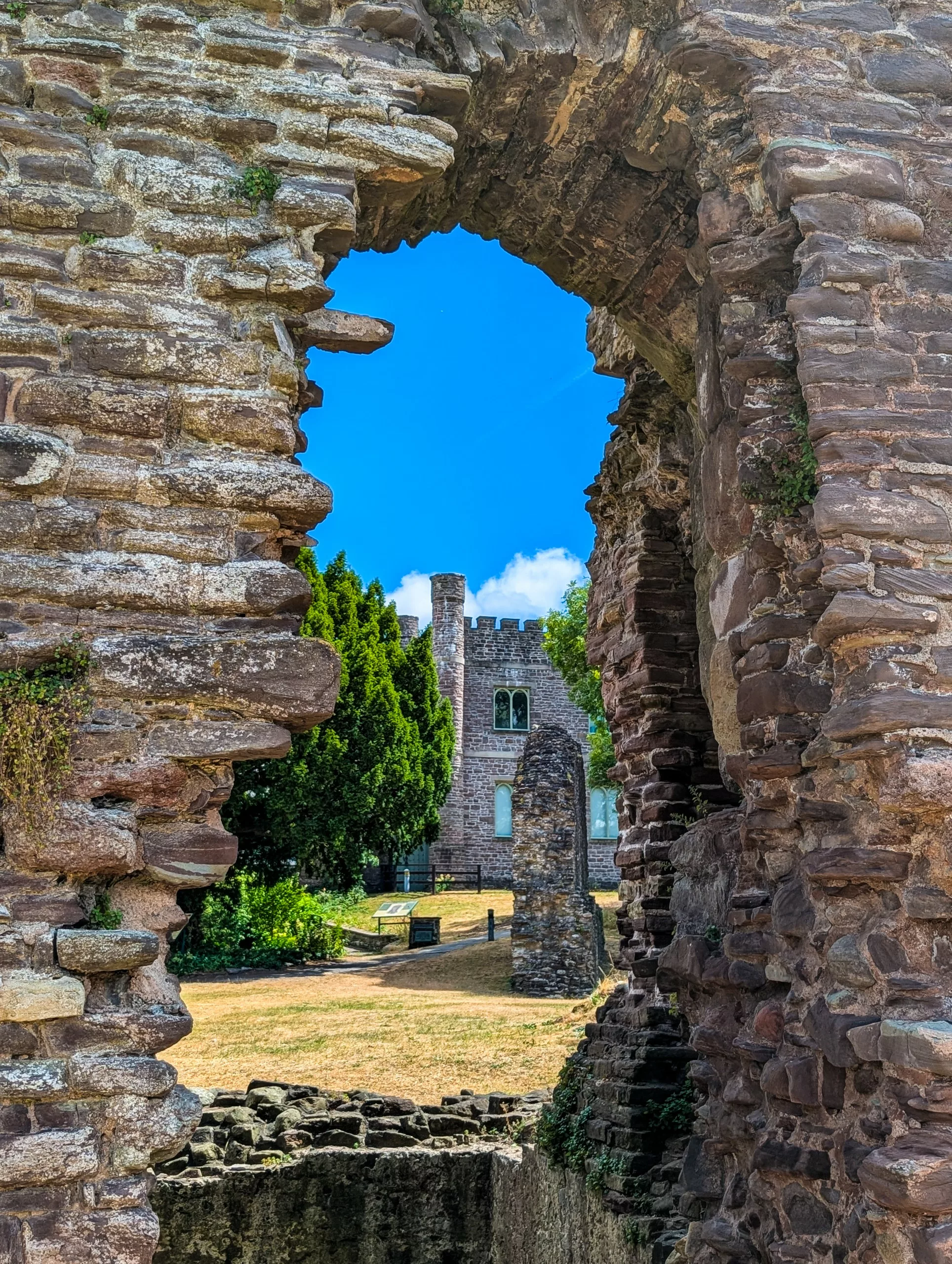 Un bâtiment en pierre doté d'une tour crénelée, aperçu à travers une ouverture irrégulière dans un mur de pierre en ruines
