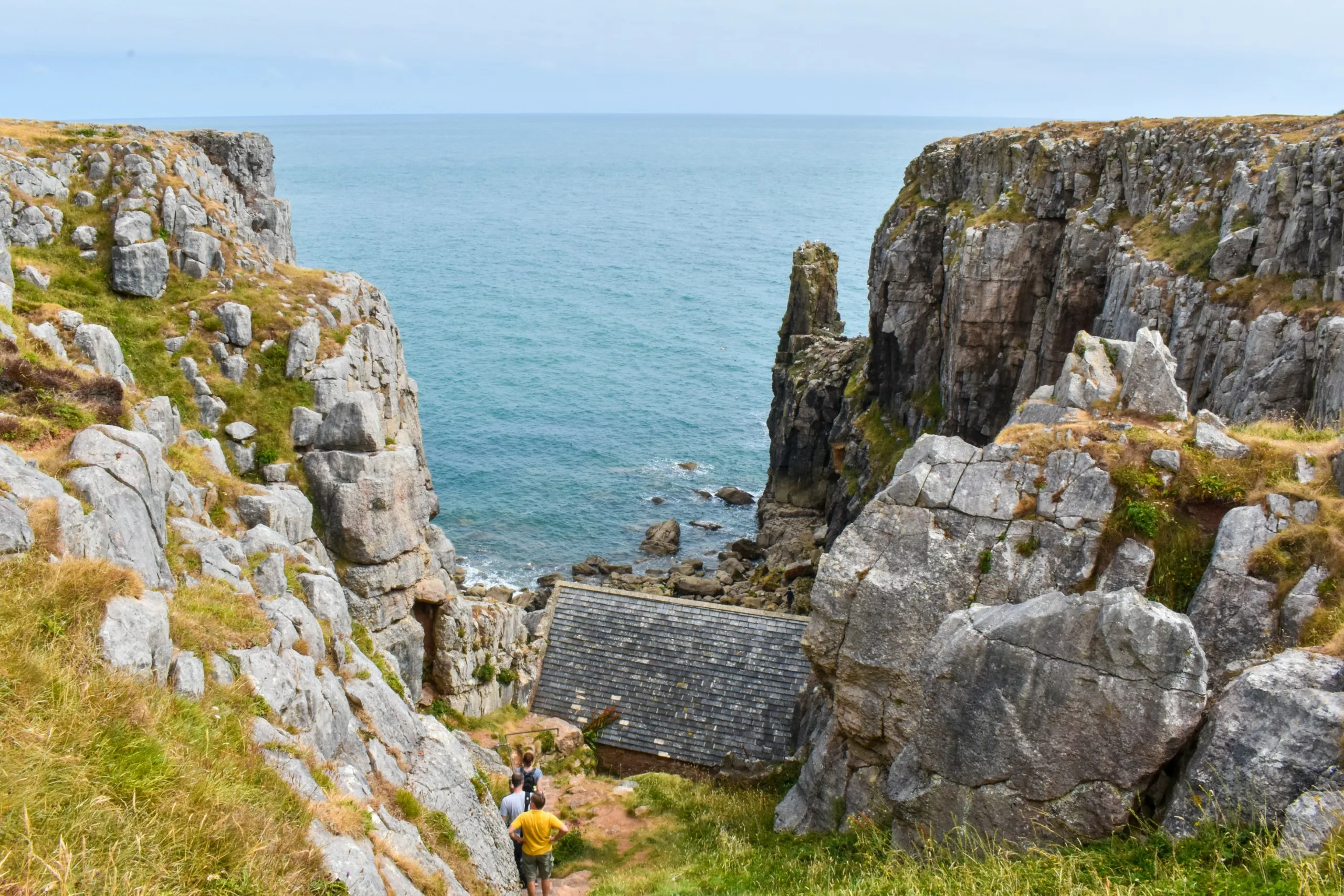 Trois personnes empruntent un sentier entre des falaises rocheuses en direction d'un petit bâtiment en pierre surplombant la mer plus bas.
