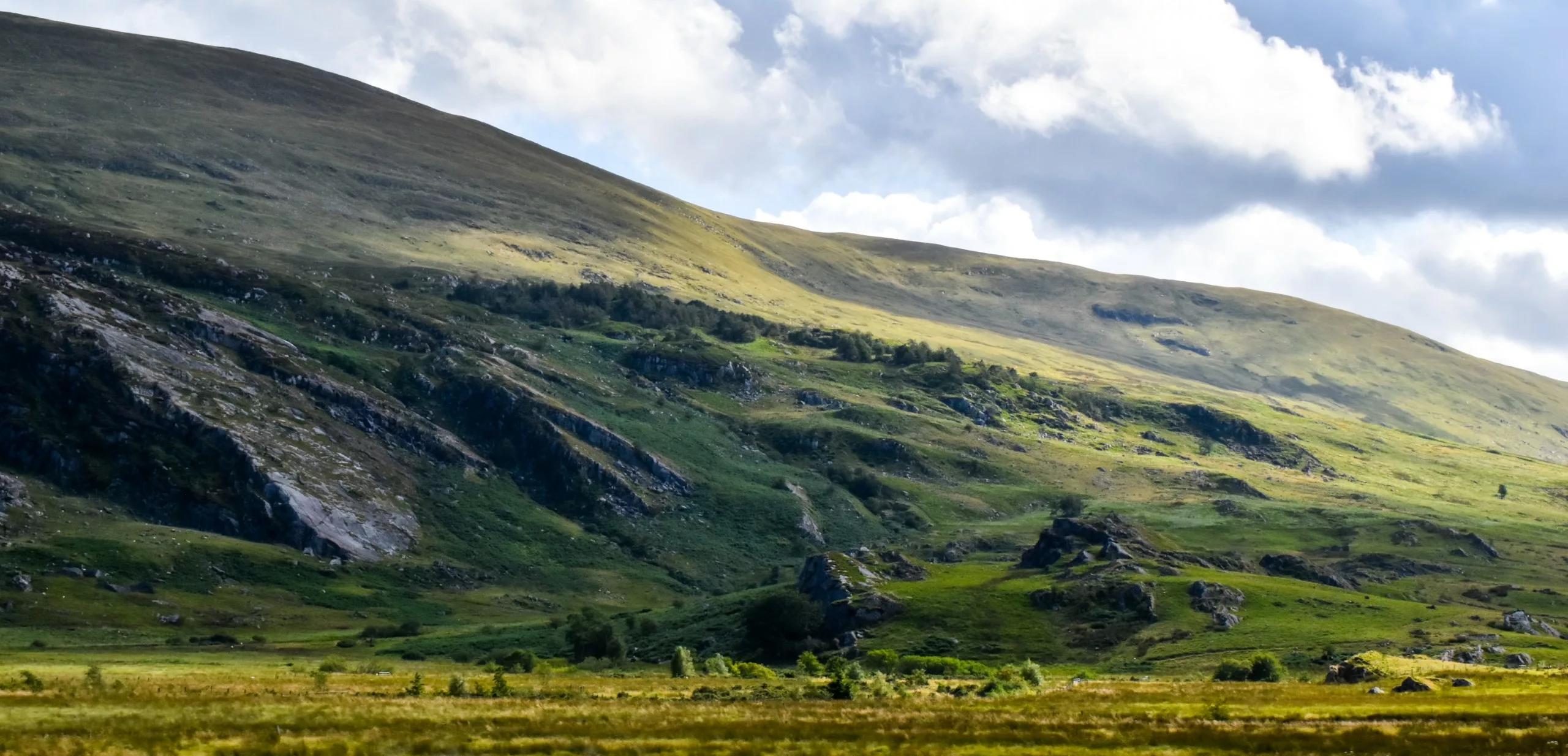 Une vaste colline verdoyante aux pentes rocheuses, sous un ciel nuageux, sur la route au Pays de Galles.