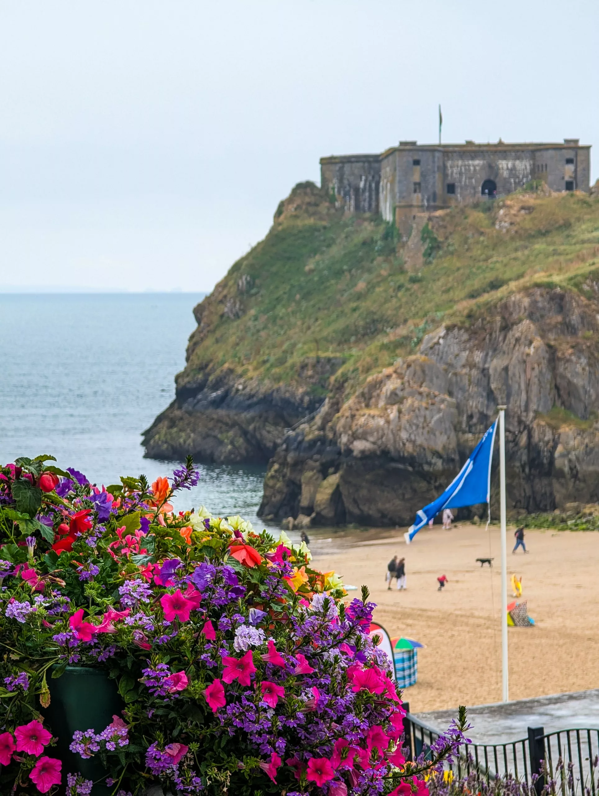Une forteresse en pierre perchée au sommet d'une falaise côtière surplombant une plage de sable, avec un drapeau bleu au premier plan et une jardinière aux couleurs vives, remplie de fleurs roses et violettes, en bas à gauche.