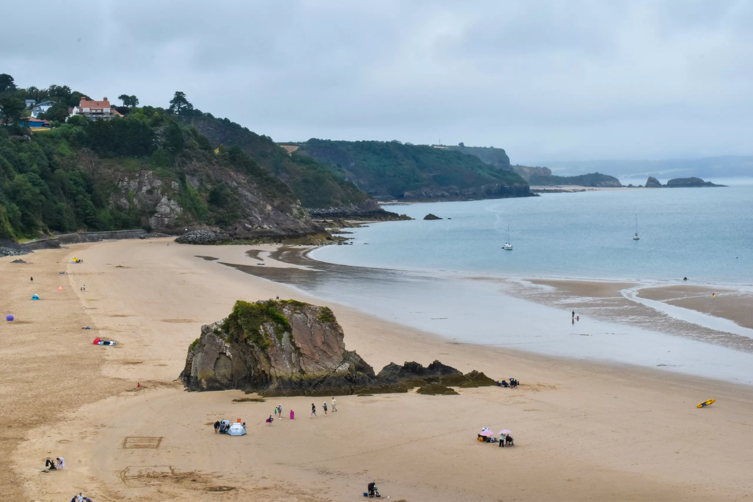 Une crique de plage de sable à Tenby, avec une grande formation rocheuse, quelques baigneurs éparpillés et deux voiliers sur l'eau, sous un ciel nuageux.