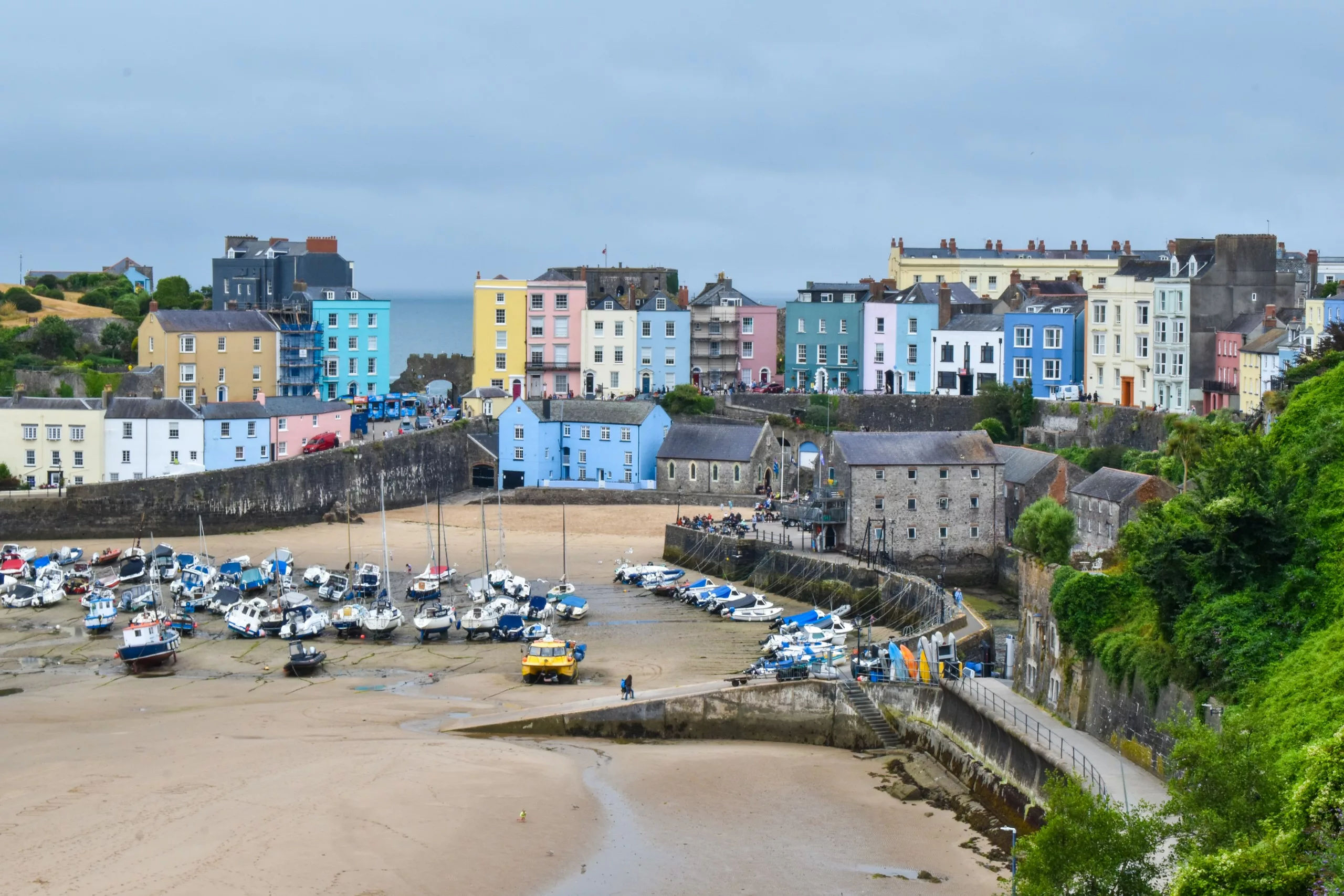 Des bâtiments colorés bordent une ville côtière du Pays de Galles, surplombant un port où de petits bateaux sont amarrés sur une plage de sable à marée basse.