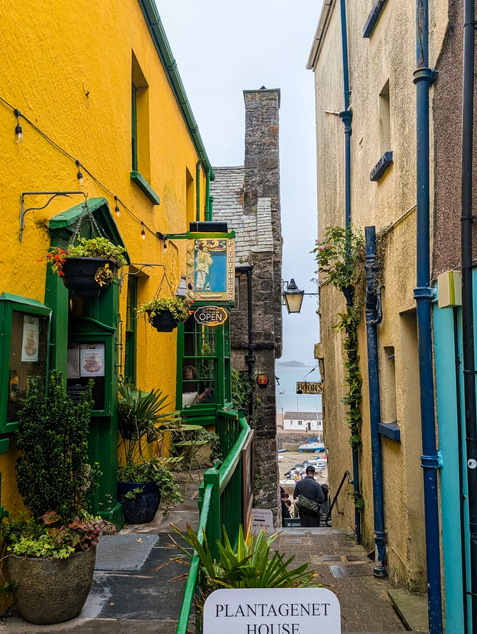 Un escalier étroit en pierre descend entre un bâtiment d'un jaune éclatant et un autre d'un beige clair en direction d'un port, avec, au premier plan, une enseigne suspendue indiquant « Plantagenet House ».