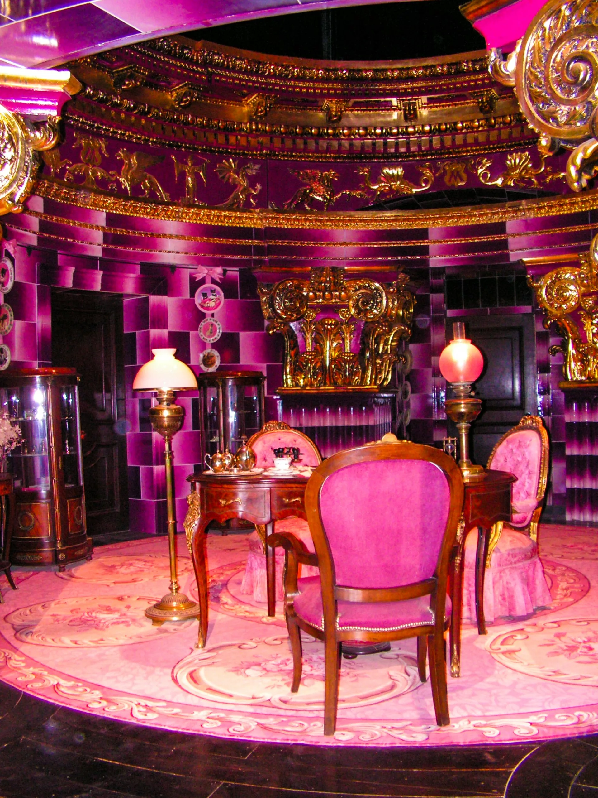 Une pièce somptueuse avec une table ronde entourée de chaises rembourrées roses posées sur un tapis à motifs floraux, le tout devant des murs recouverts de carreaux violet foncé et ornés de moulures dorées.